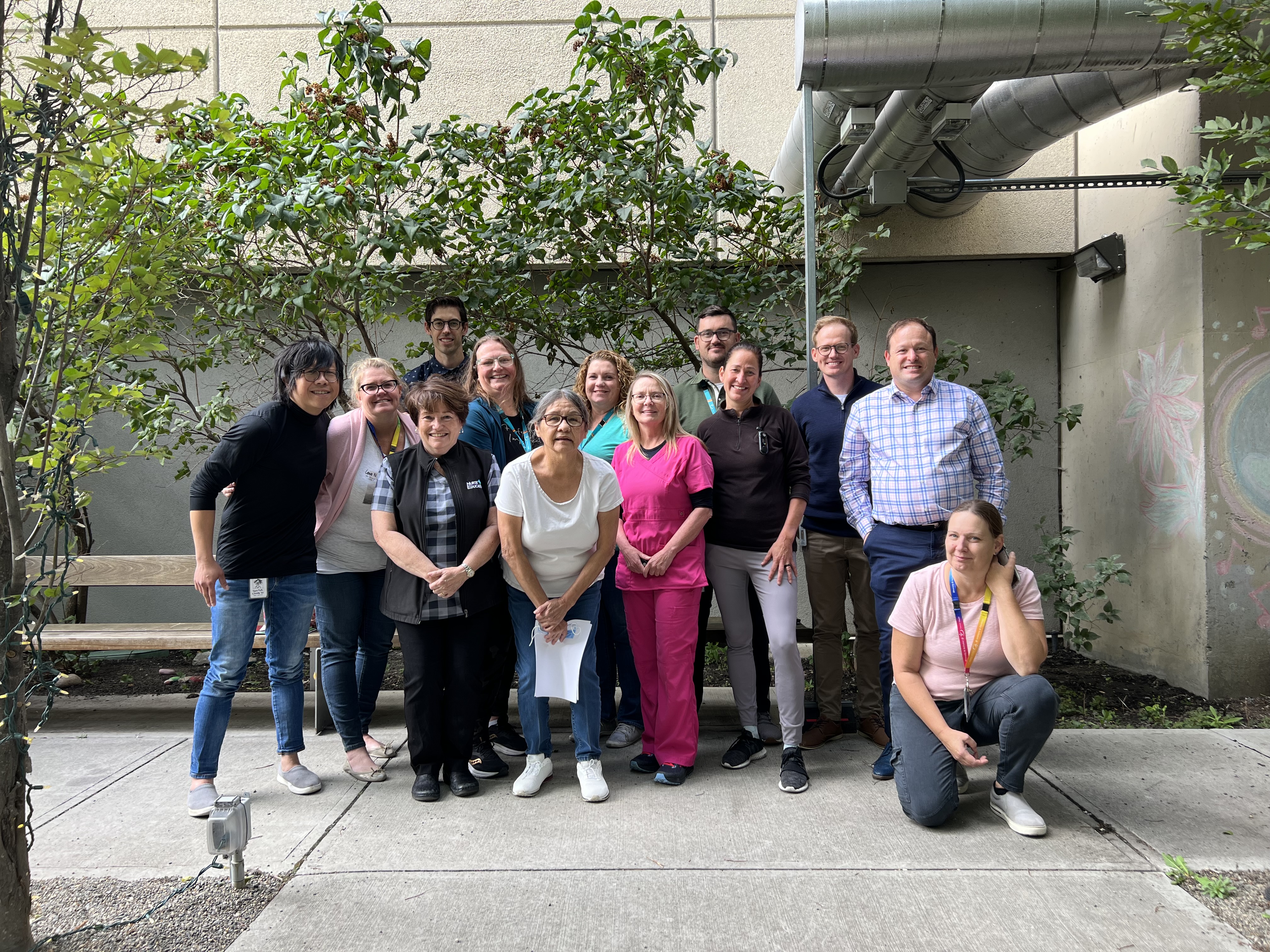 A group of smiling health care workers stand together in a group outside a hospital surrounded by small trees. 