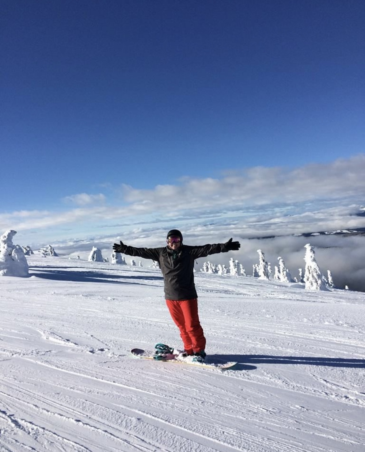 A woman wearing a helmet and bright red snow pants holds out her arms while snowboarding on the top of the mountain while clouds are surrounding the area