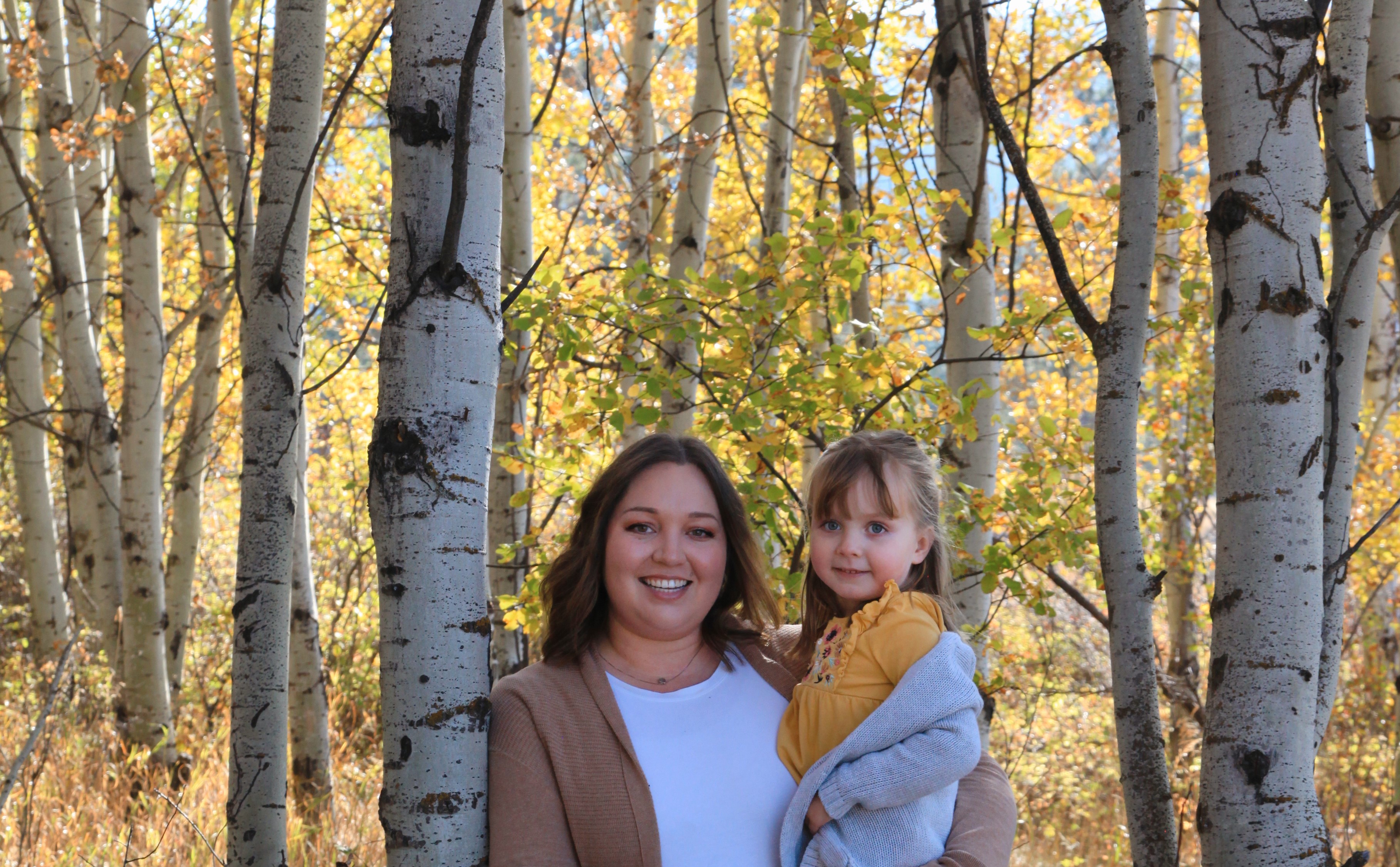 A mother and daughter smiles while standing among many trees on a sunny day