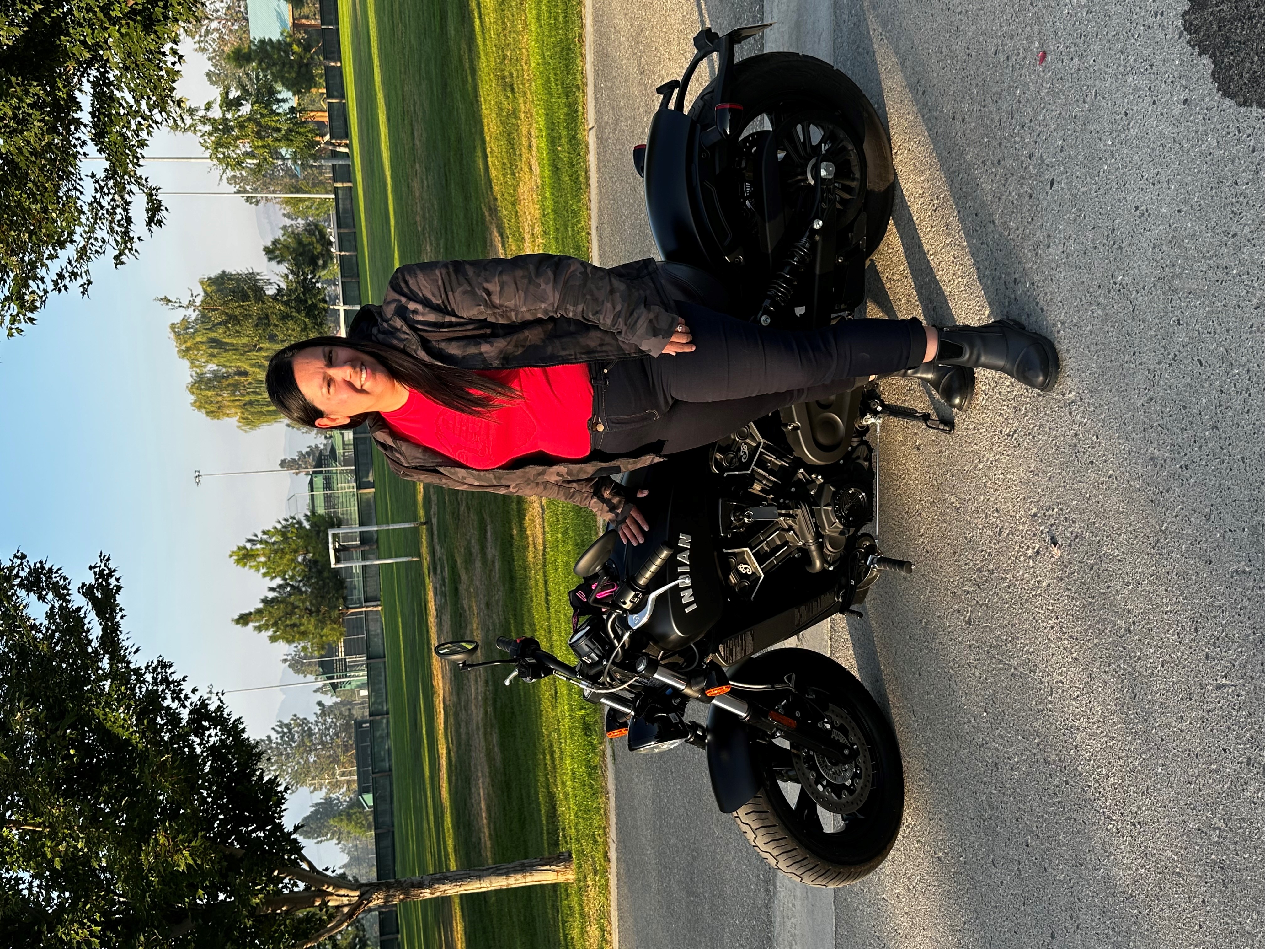 A woman with long brown hair in a bright red shirt leans on an upright motorcycle posed in front of a soccer field