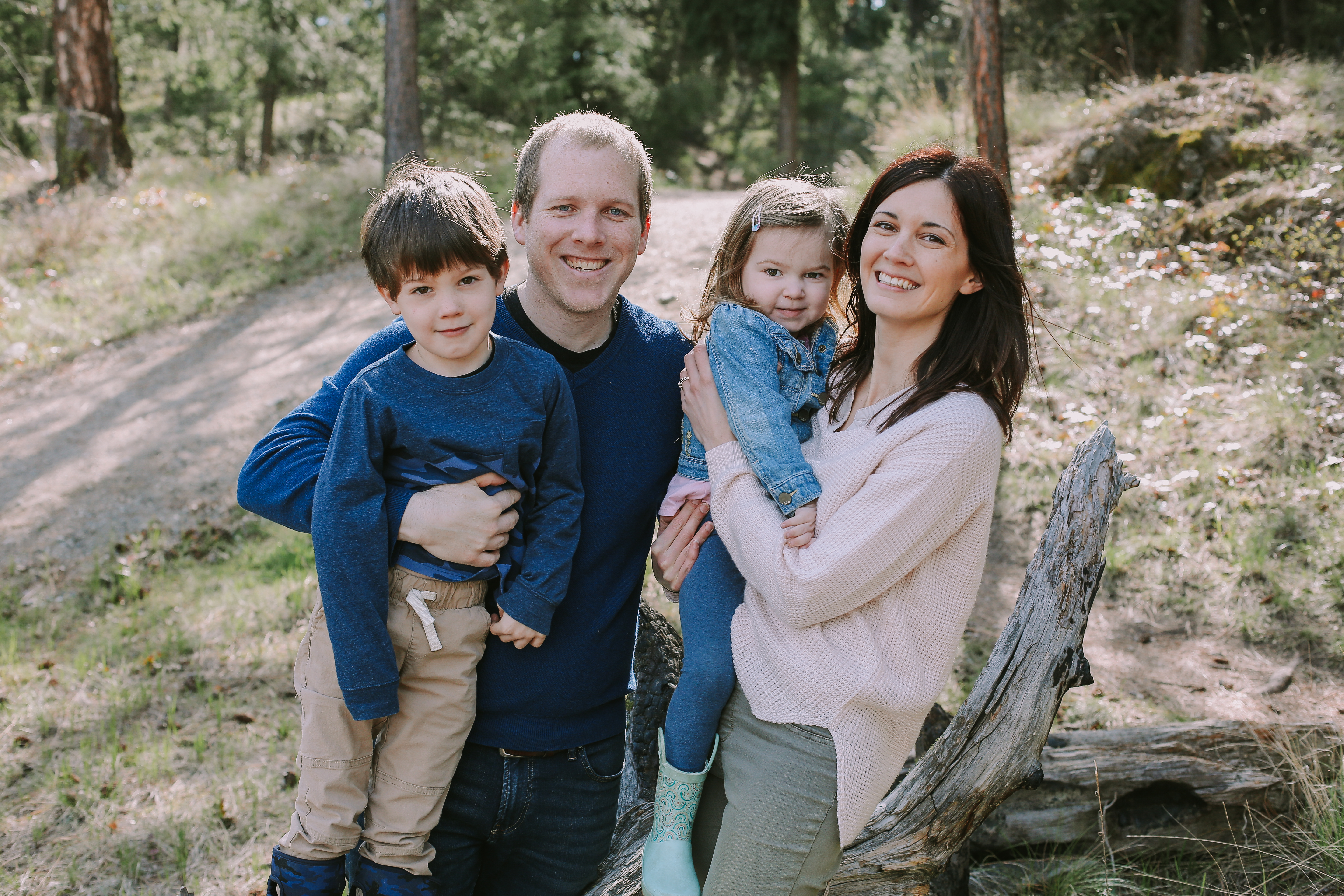 Young family with dad holding boy son and mom holding girl daughter, wearing blue and beige, standing by fallen tree and trail in forest