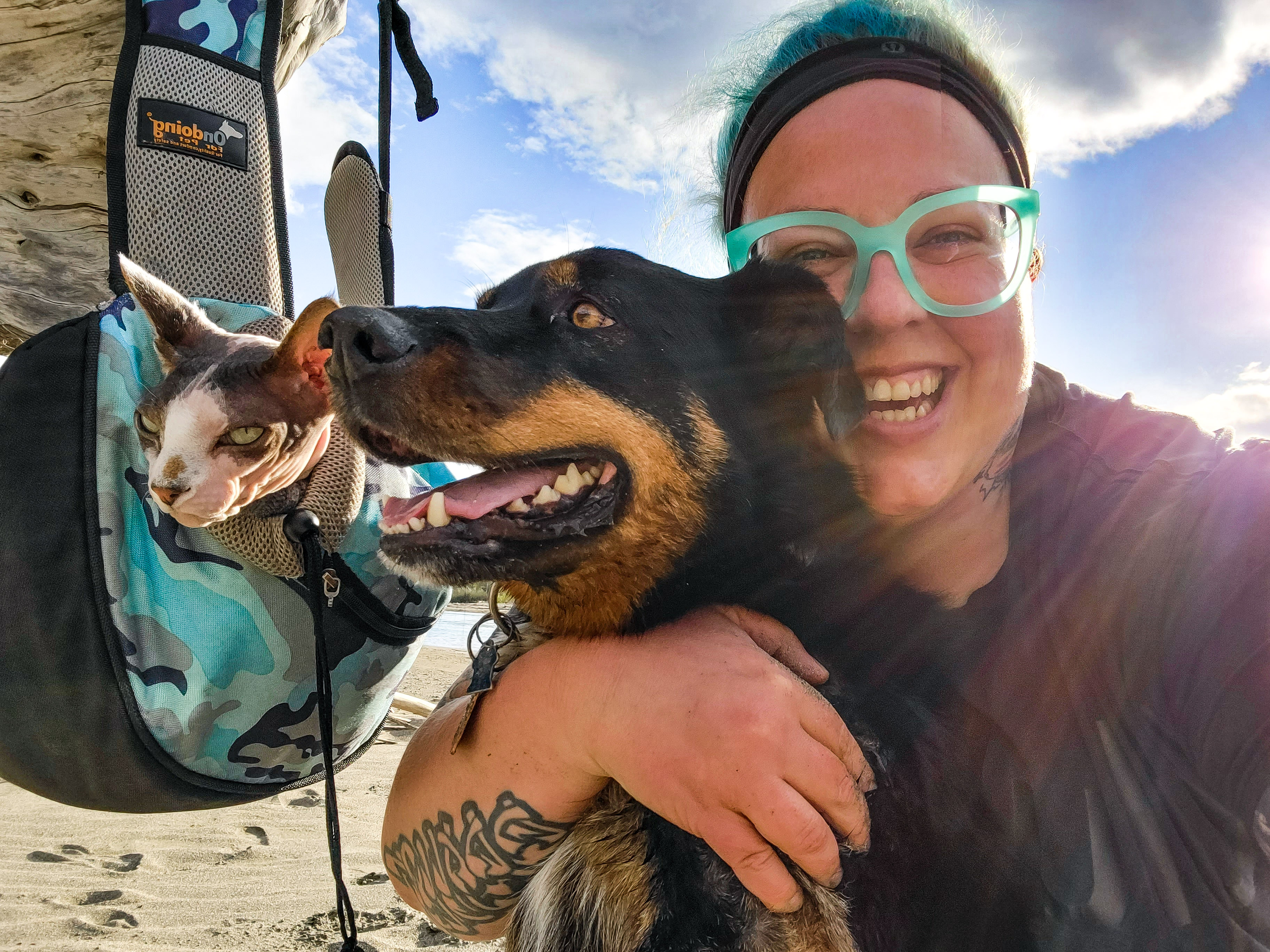 smiling blonde woman with glasses hugging her black and beige dog and bald cat on a hike