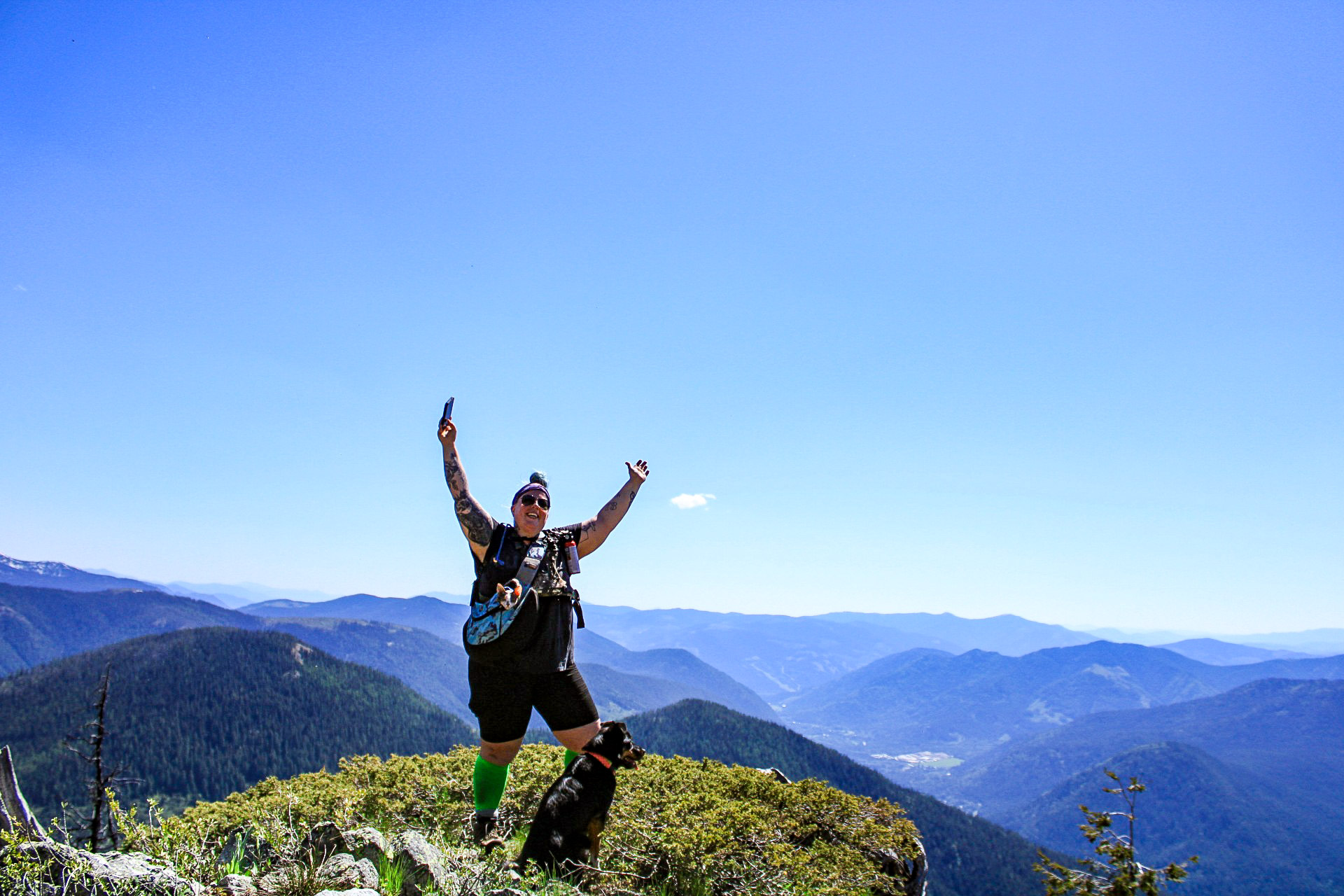 A woman standing on a mountain carrying a cat in a backpack with a black dog standing next to her.