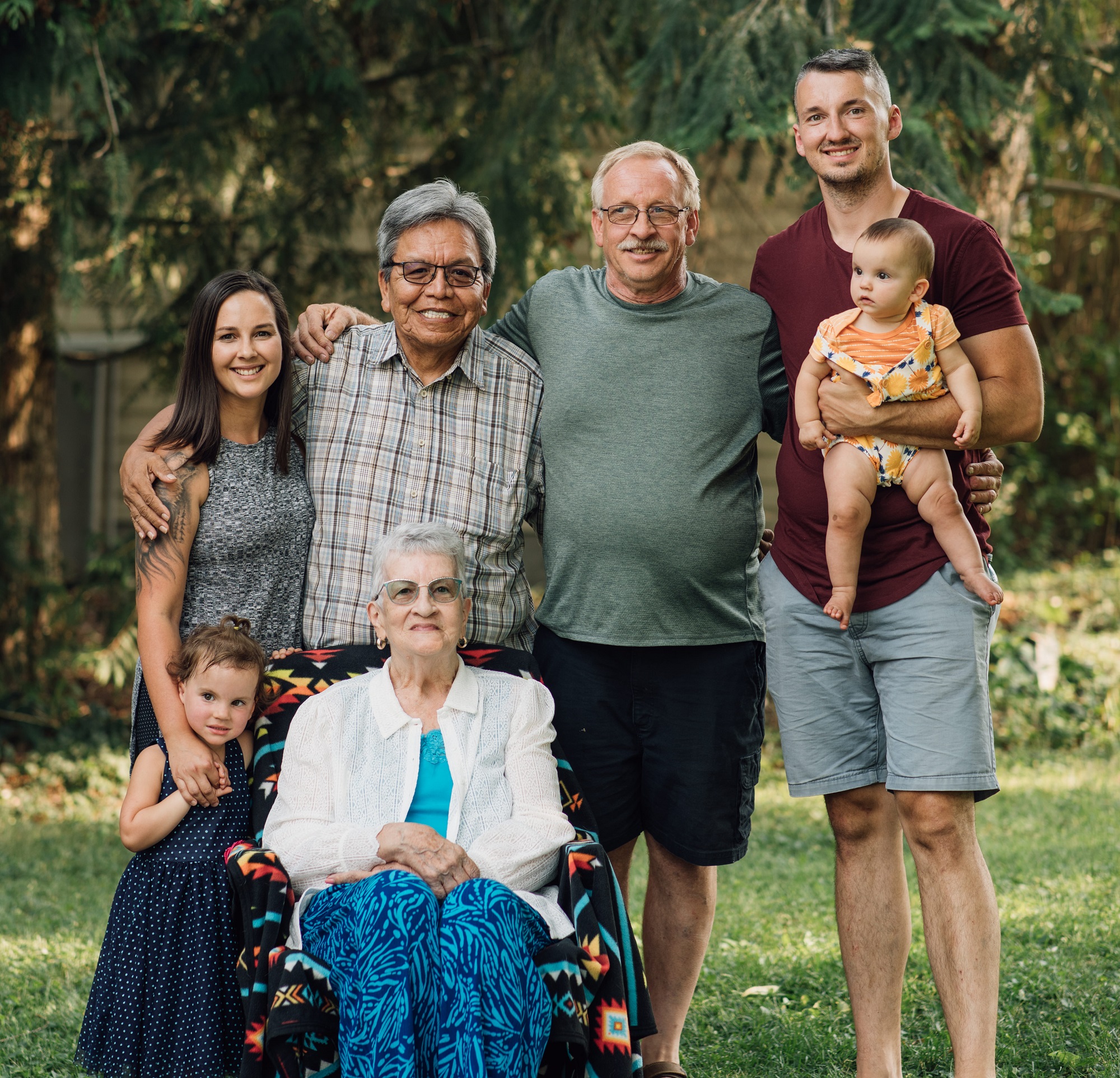 outdoor Intergenerational family photoshoot for 7 placed in two rows. 5 in the back row all standing shortest to tallest to include (left to right) one adult female, two elderly males and 1 adult male holding a toddler. Front row a small female child standing next to a seated elderly woman. All smiling. 
