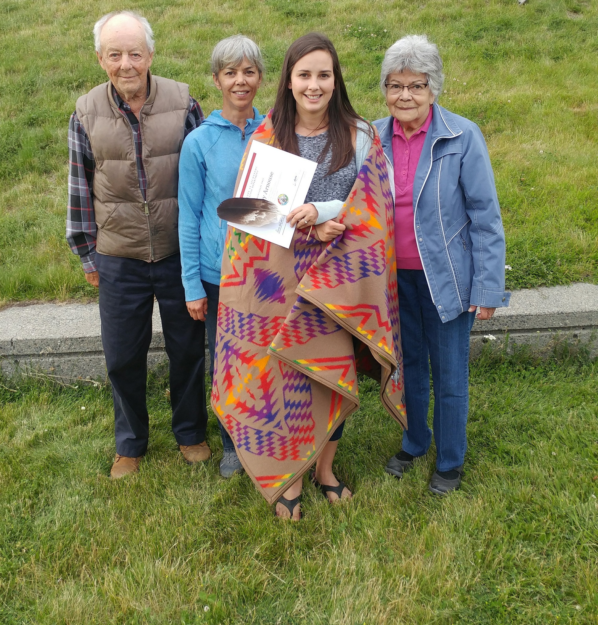 Indigenous woman wrapped in a ceremonial Pendleton blanket holding a feather and certificate with an elderly male and female on her left and an elderly female on her right. All smiling. 