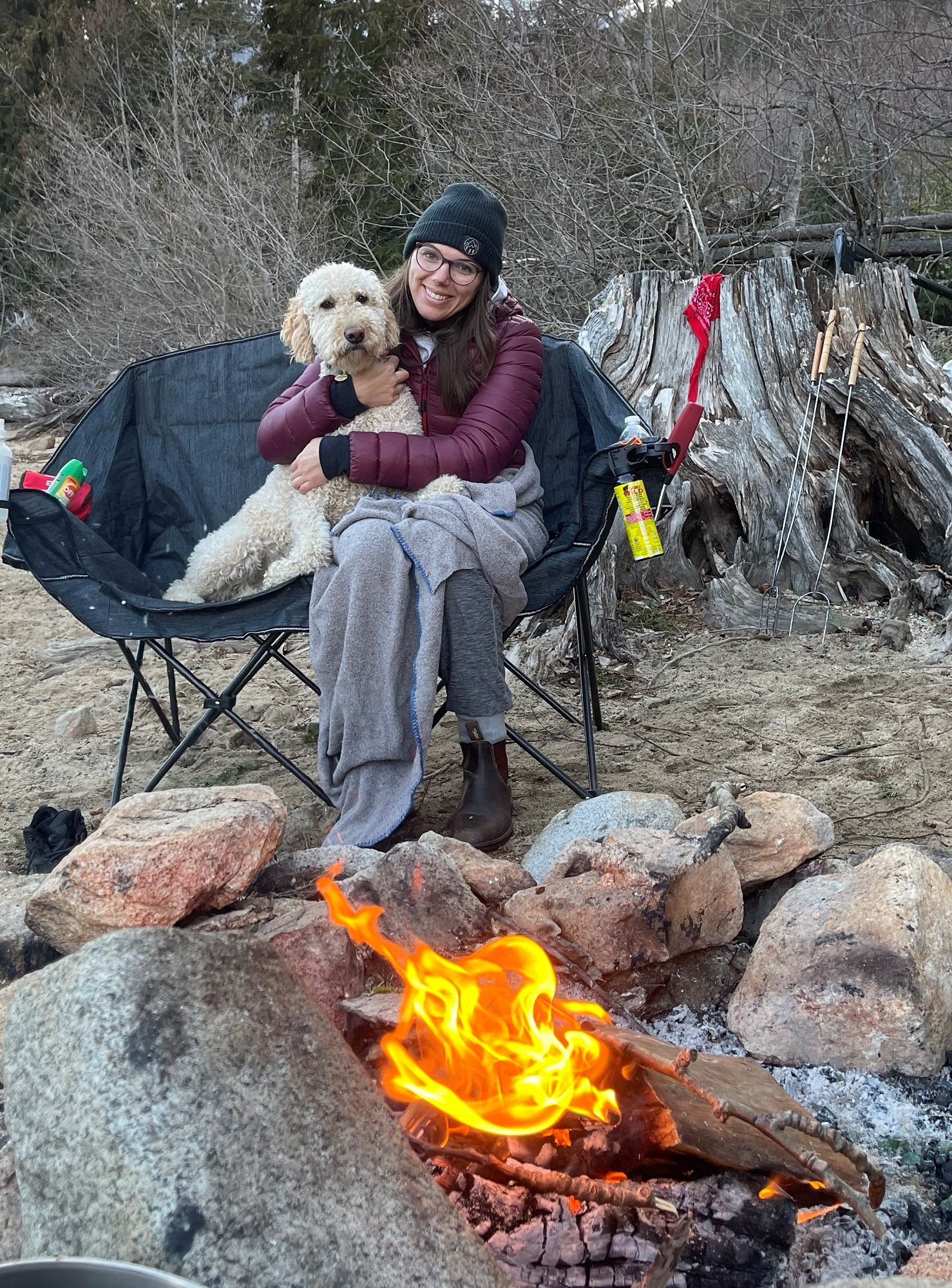 A person sitting in front of a fire in a double-wide camp chair hugging a white fluffy dog.