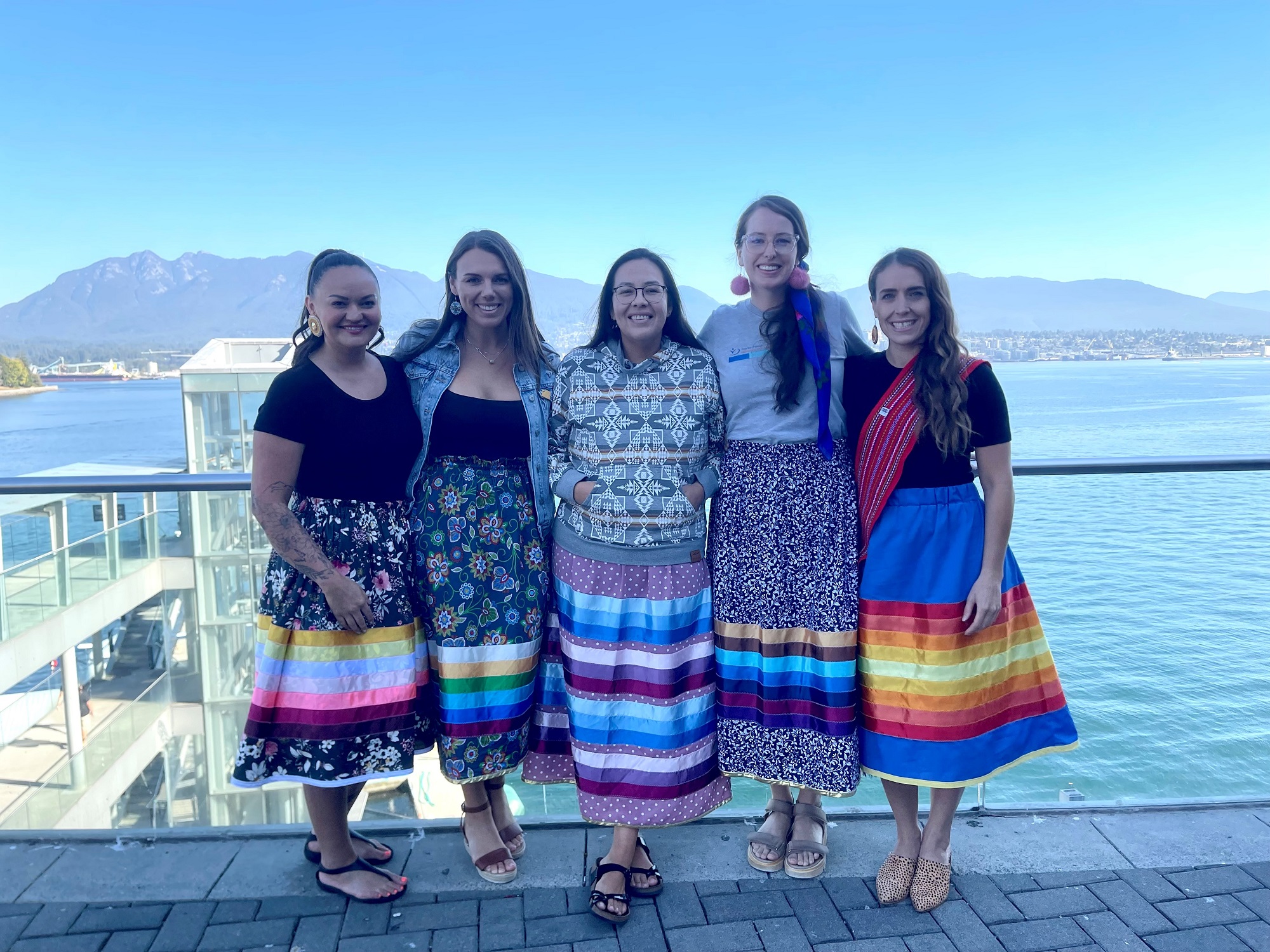 A group of women in ribbon skirts posing for a picture on a balcony with a view of the ocean and mountains behind them.
