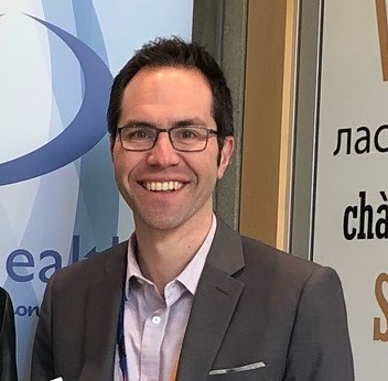 A smiling man with short black hair and black oval glasses wears a white shirt and grey jacket standing in front of an interior health logo