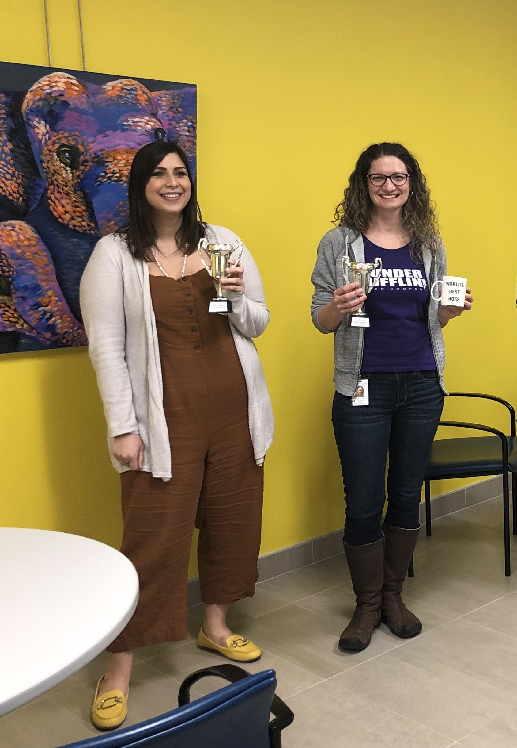 Two women standing together, each holding a trophy, one woman is holding a trophy and a coffee mug that says world’s best boss