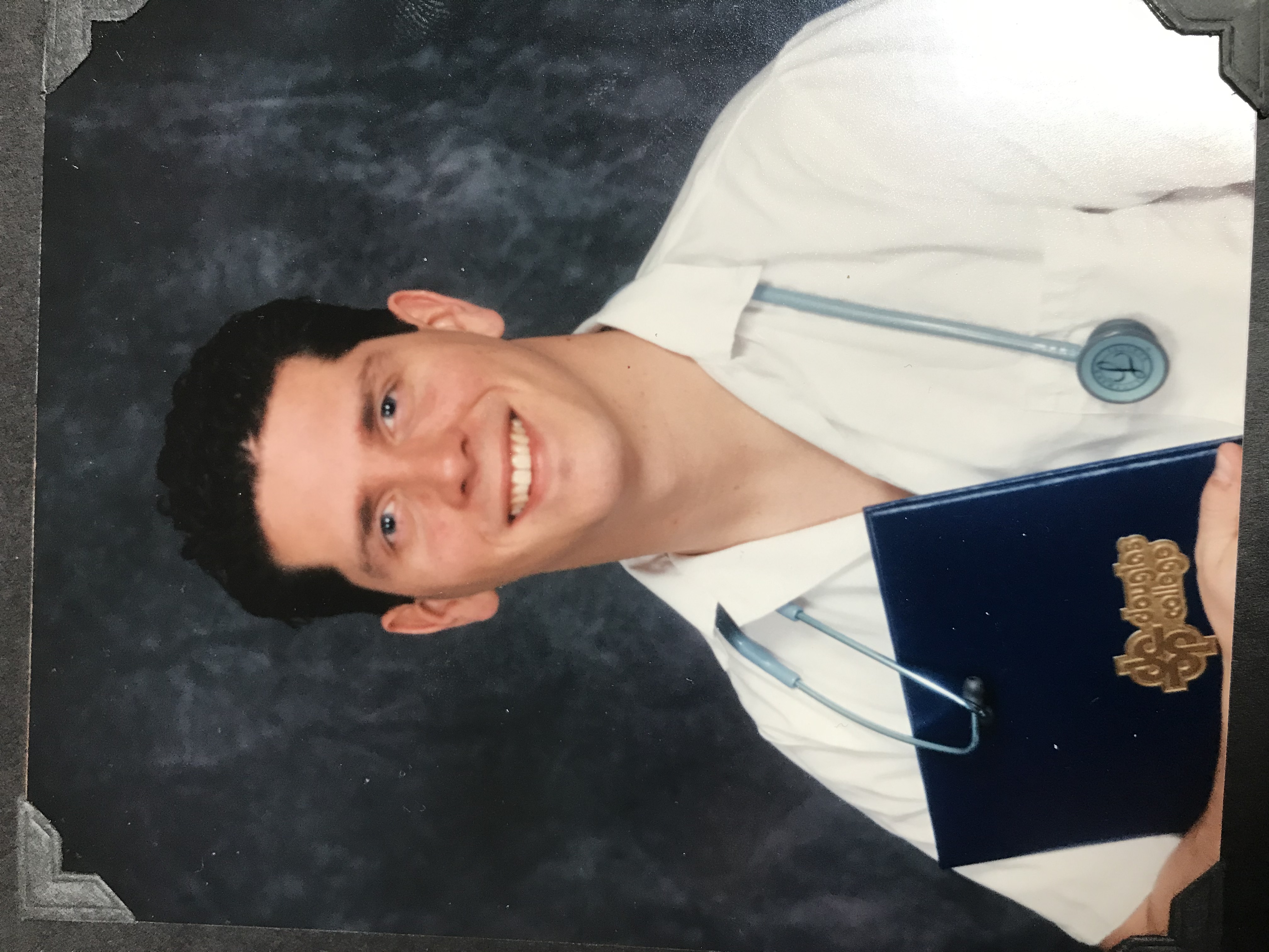 A man with short brown hair wearing a stethoscope and holding a degree certificate