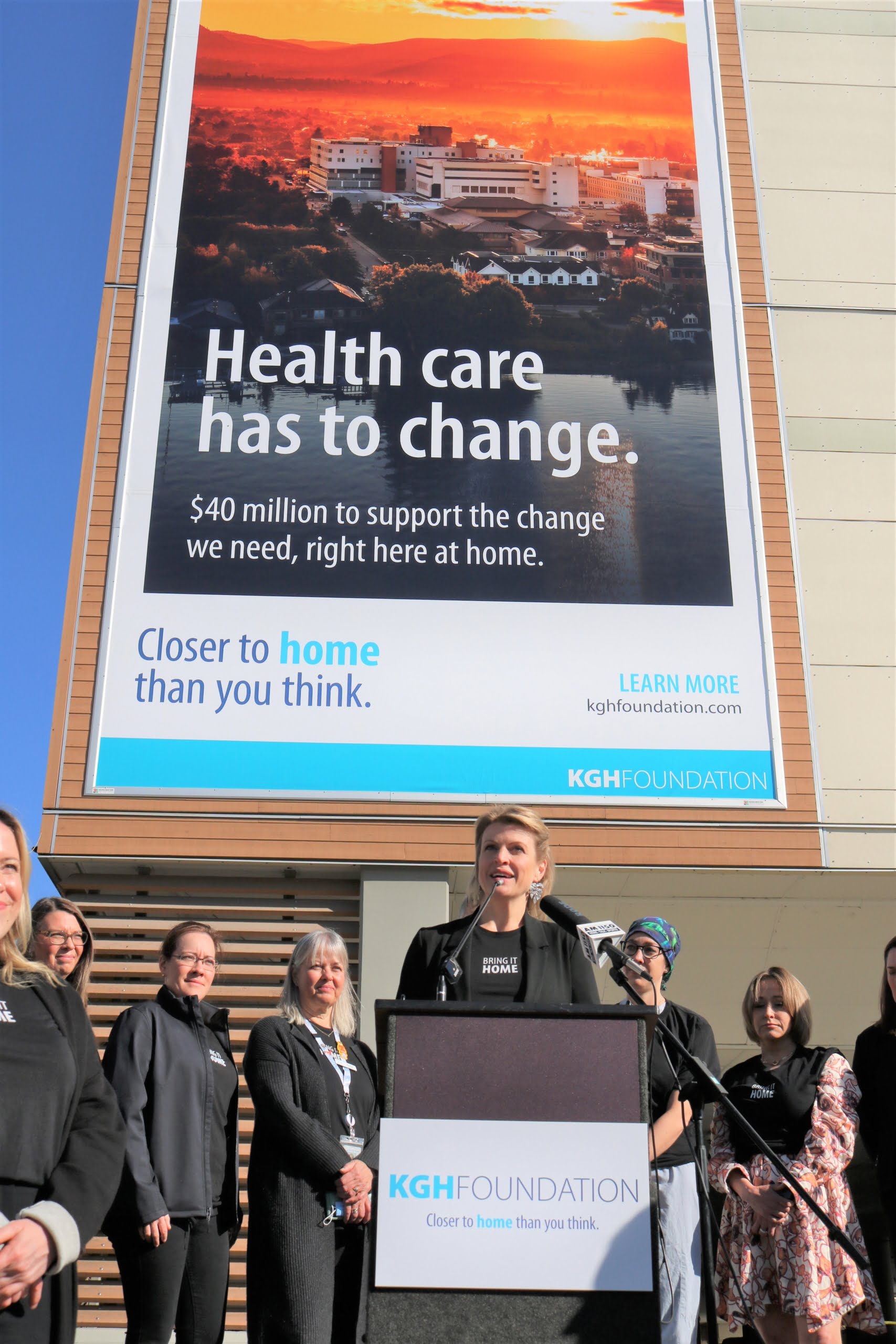 Person speaking at a podium with an audience in the foreground, with a large banner in the background displaying an advertisement for KGH Foundation that reads "Health care closer to home. $40 million to support the change we need, right here at home.