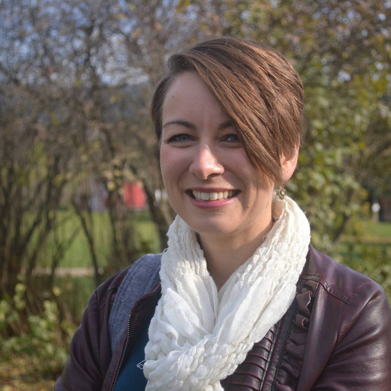 A smiling woman with brown hair tied back into a bun wears a white scarf and black leather jacket stands in front of blurred trees