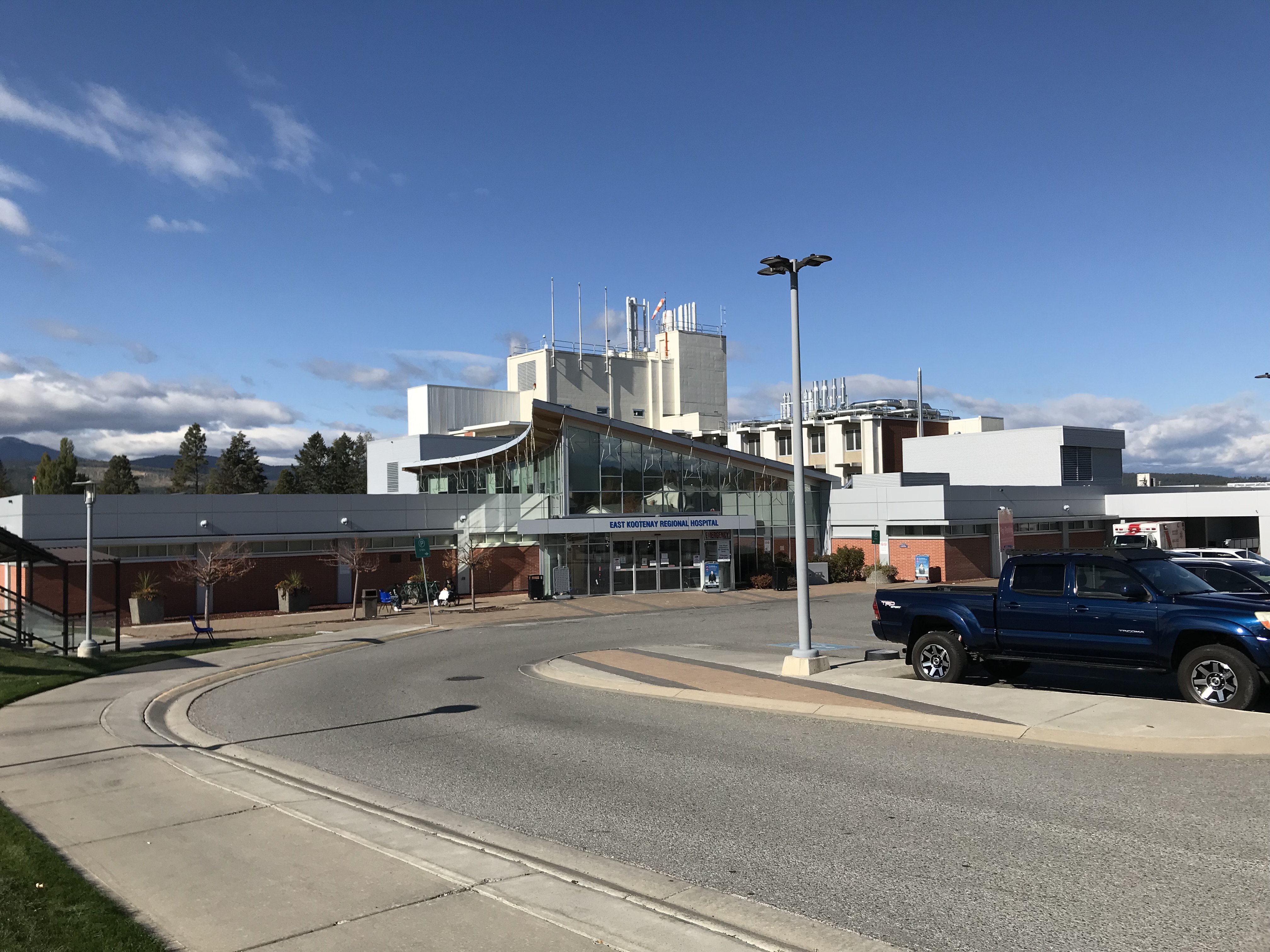 Exterior view of the East Kootenay Regional Hospital featuring modern buildings with a clear glass entrance and distinctive white structures on the roof, under a partly cloudy sky. There's a driveway in the foreground with parked vehicles.