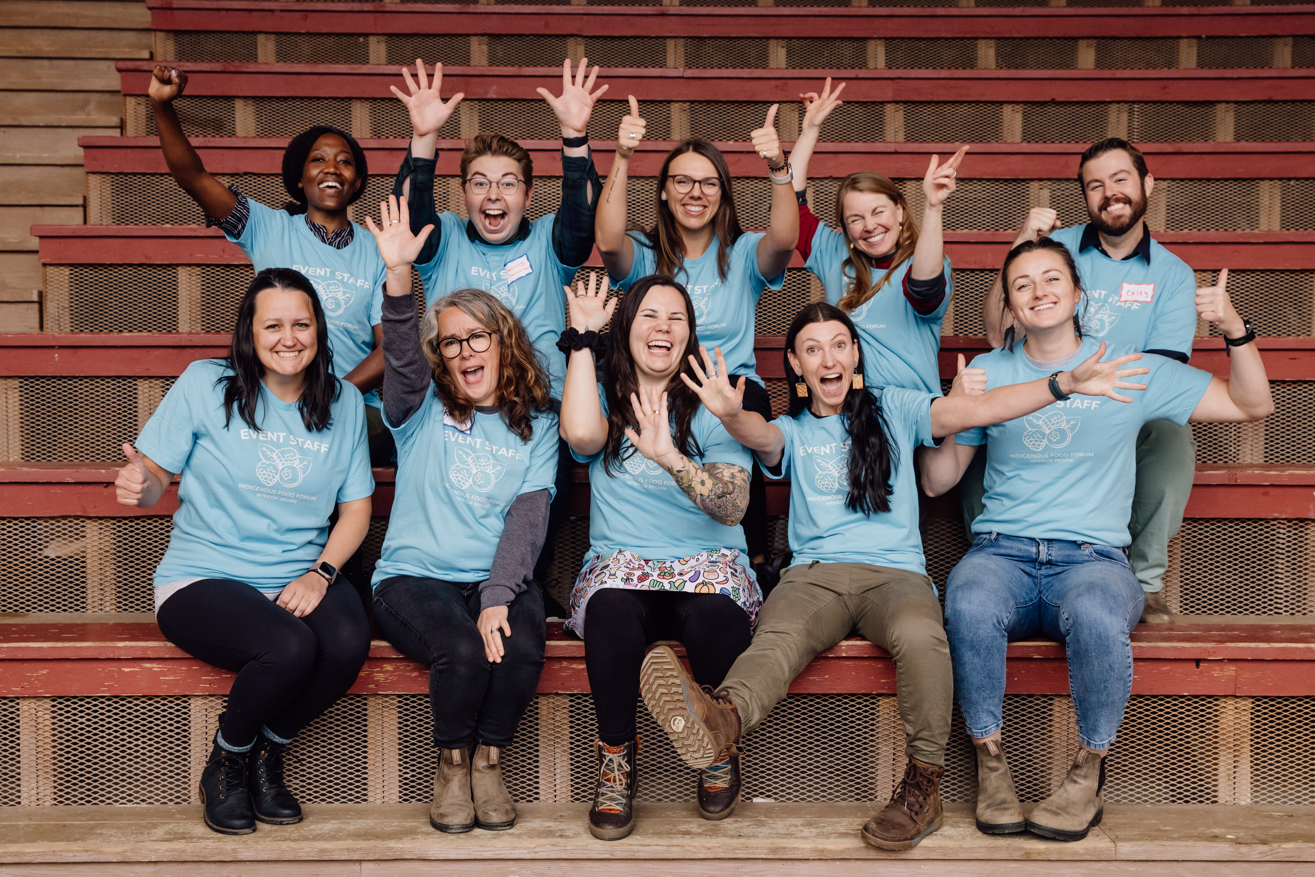  A group of people in blue shirts that say "event staff" have fun posing for a picture while seated on wooden bleachers.