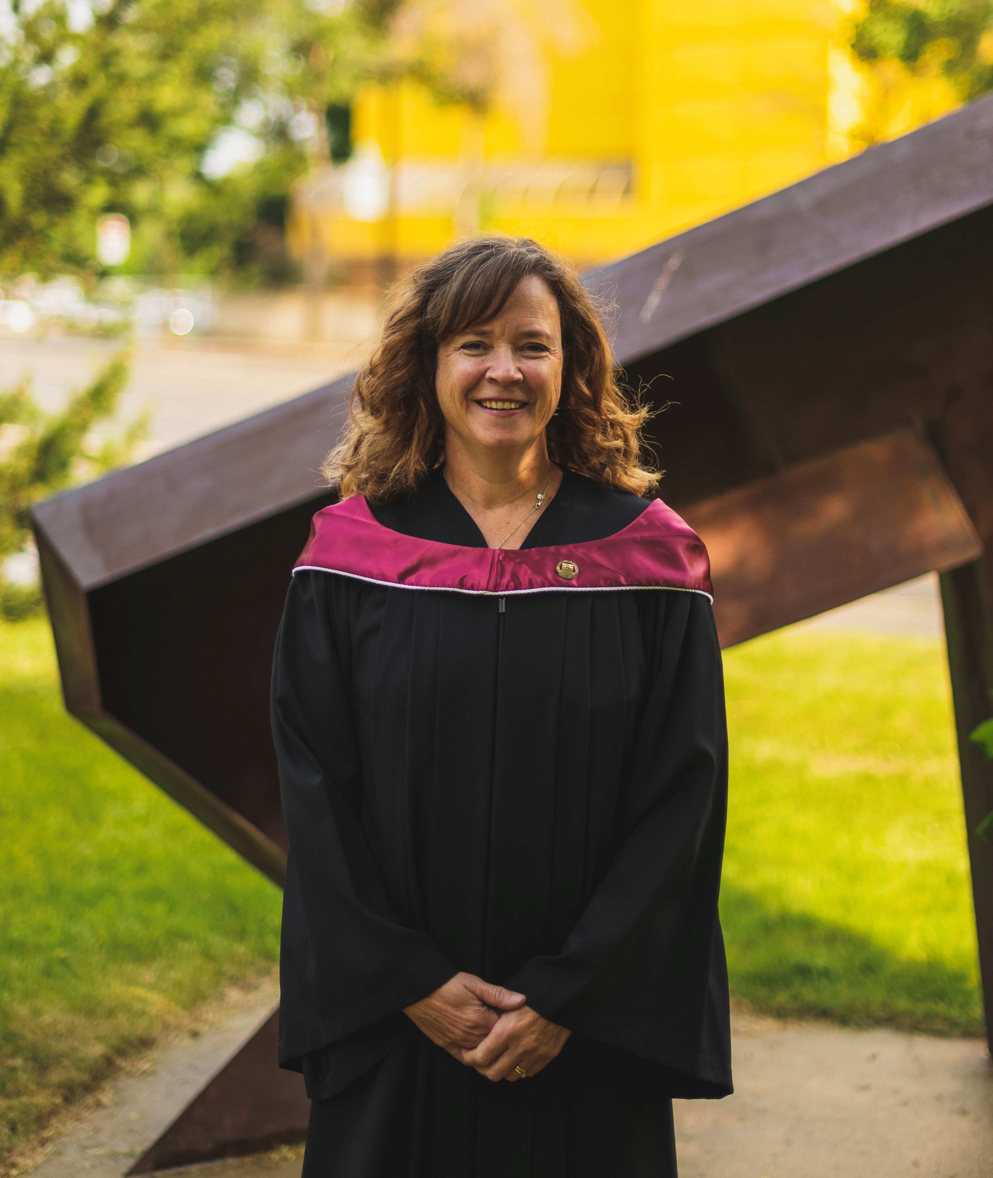 A woman with shoulder length curly brown hair and wearing a black robe and pink collar graduation gown smiles in front of a sculpture