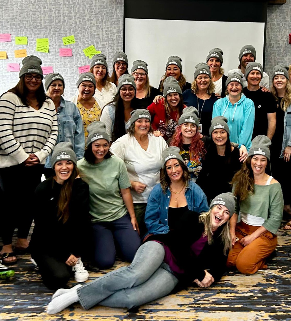 Group of people smiling in a room, many wearing gray beanies, with colorful sticky notes on the wall behind them.