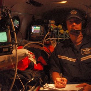 A man in a blue ball cap and short-sleeved shirt wearing a headset sits inside an airplane with a medical patient on a gurney surrounded by machines