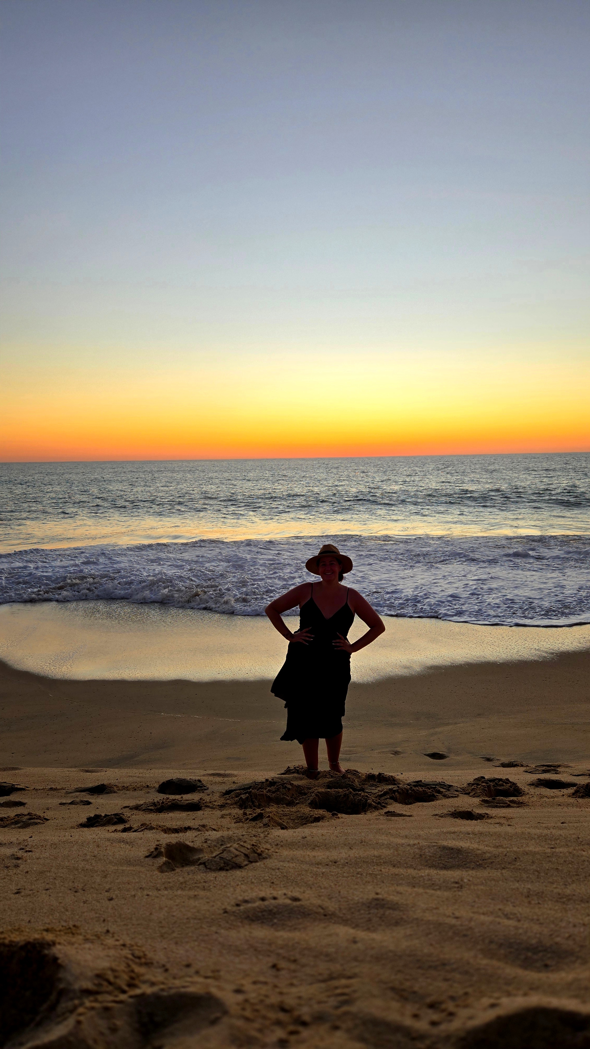 Person standing on a beach at sunset with gentle waves in the background.