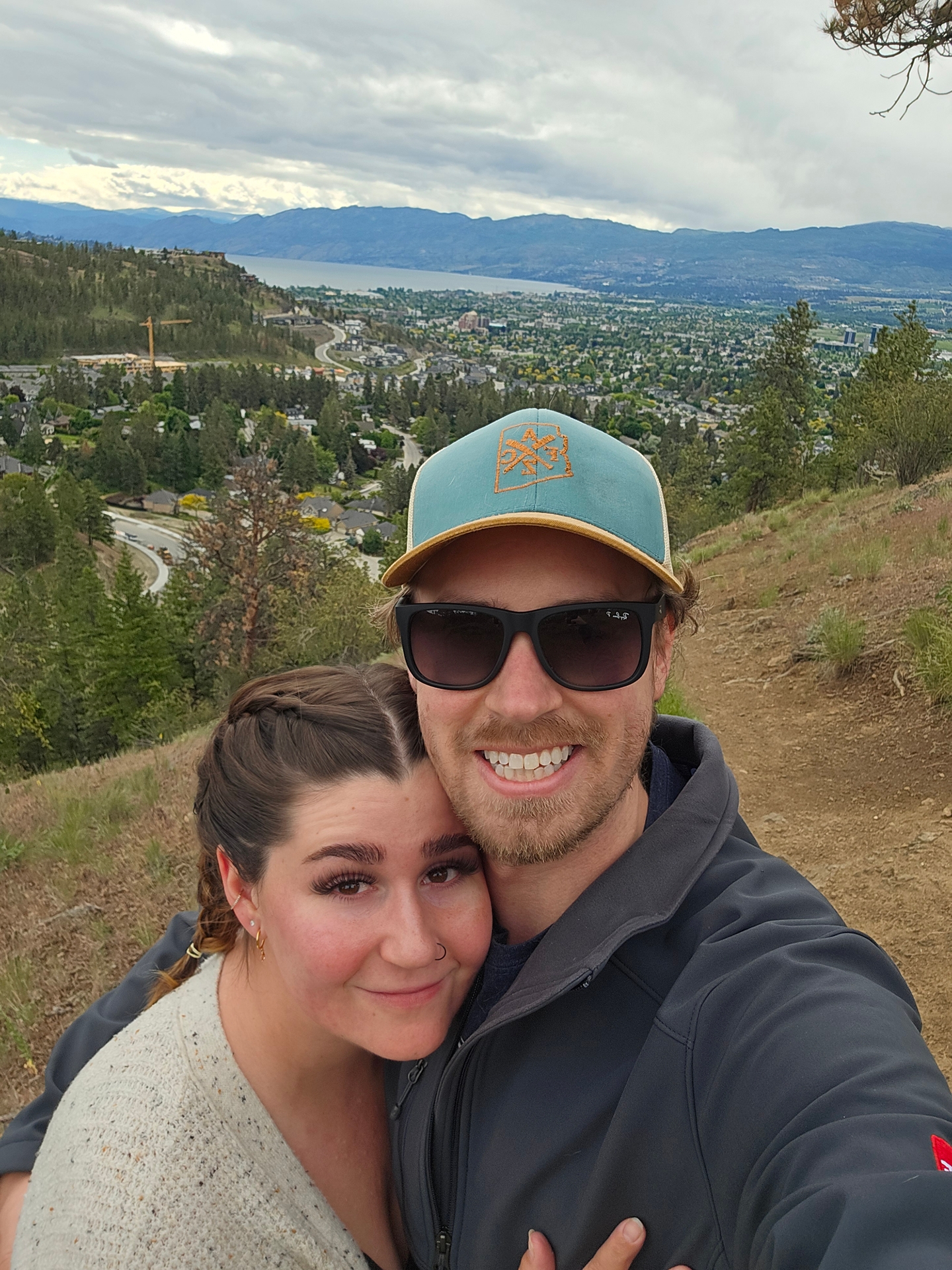 Two people smiling for a selfie with a scenic view of hills and a city in the background. One person is wearing sunglasses and a cap, and the other has their hair styled in braids.