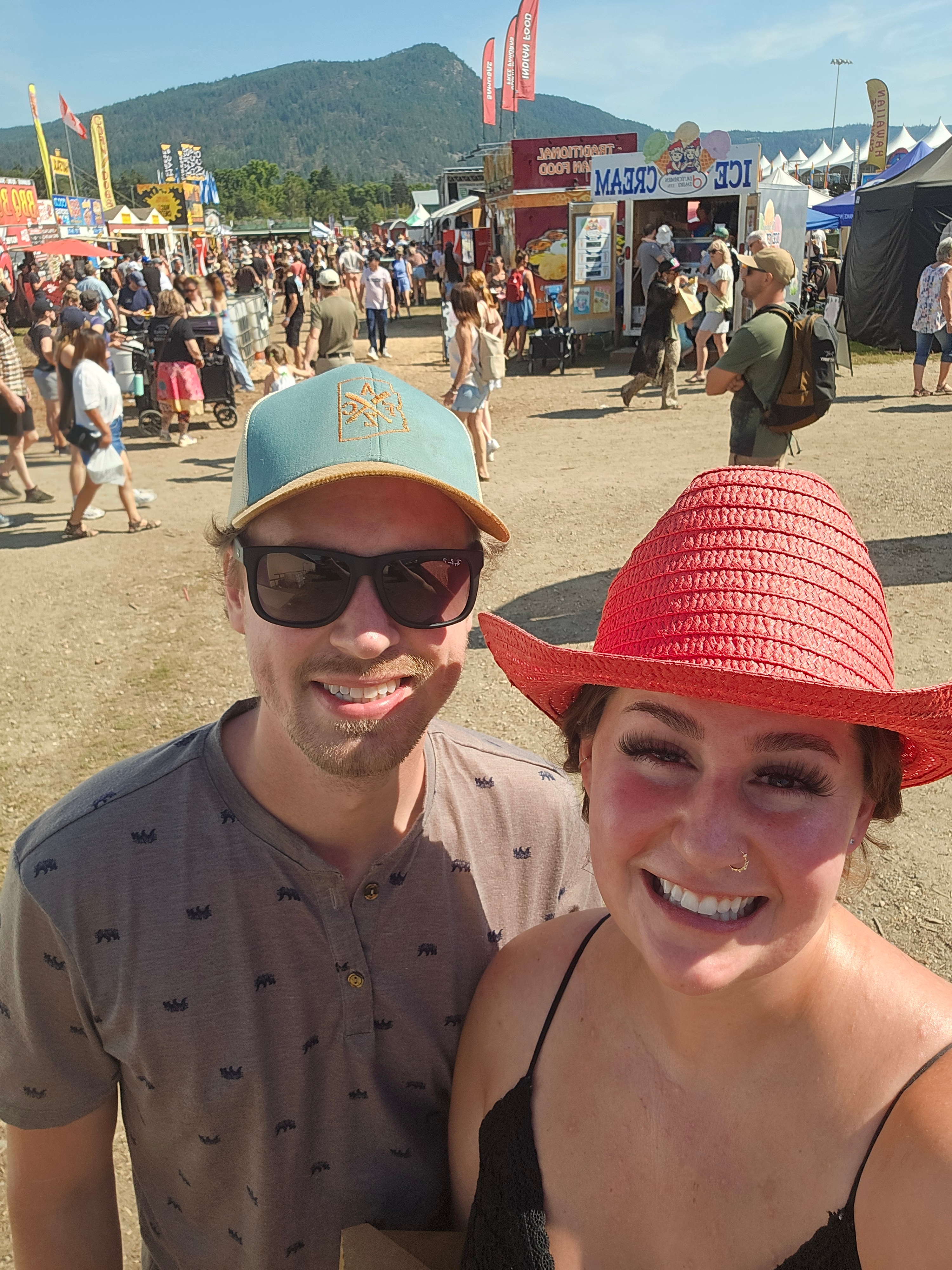 Two individuals smiling at a bustling outdoor festival with food stalls and a mountainous backdrop. One person is wearing a teal cap and sunglasses, and the other is in a red woven hat.