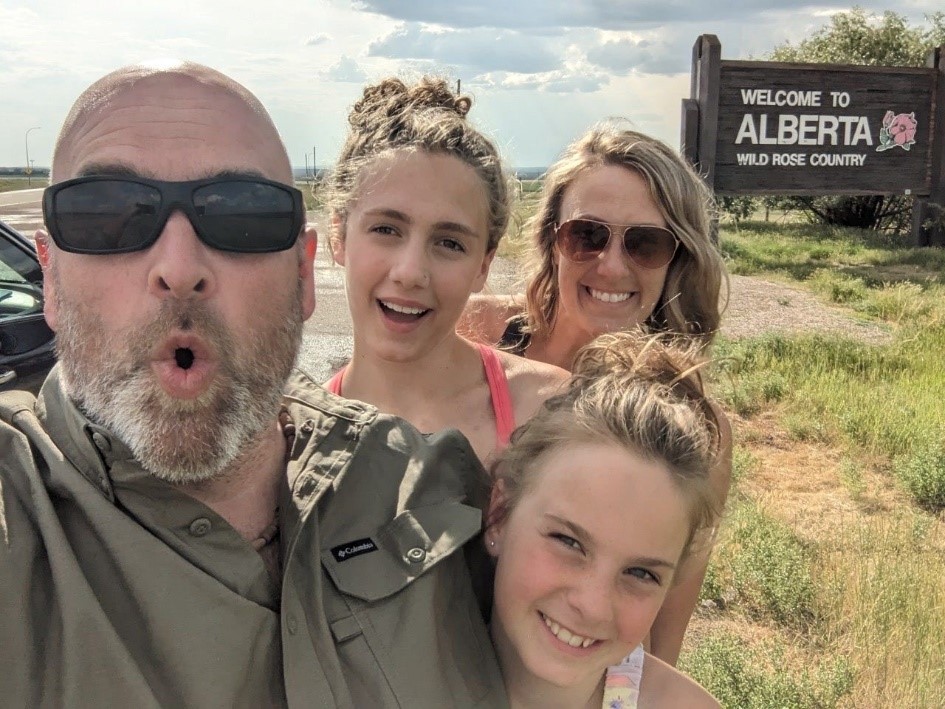 A man with a beard and sunglasses and khaki shirt makes a funny oh face as he poses with his family, two young girls with their hair in a bun and a woman with blond hair and sunglasses all posing in front of a Welcome to Alberta Wild Rose Country sign