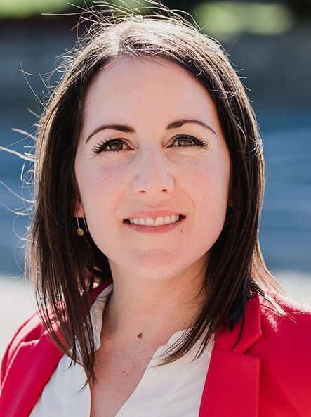 A head and shoulders photo of a smiling woman with straight brown shoulder-length hair wearing a red jacket and a white blouse