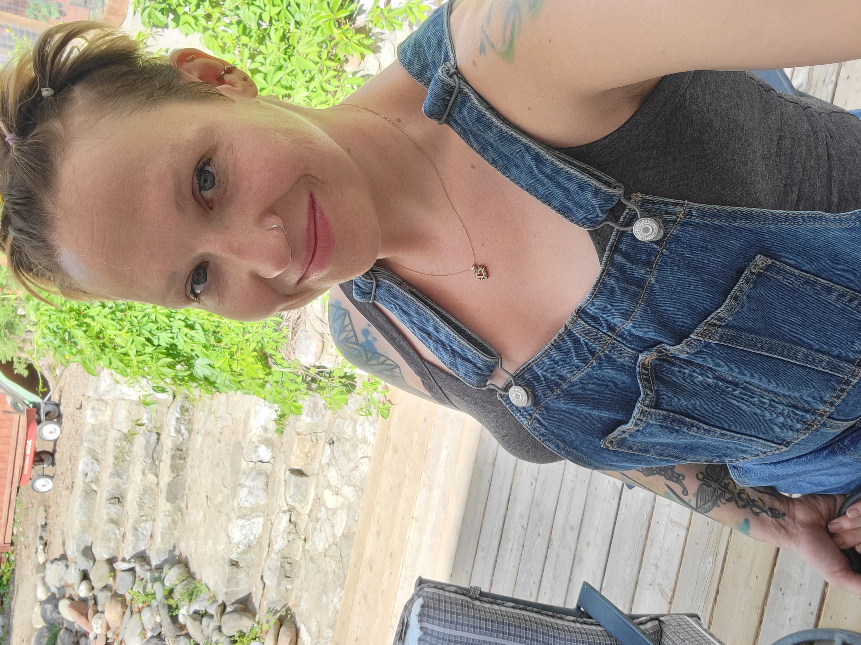 A woman with short spiky brown hair, nose ring and tattoos on her shoulders wearing jeans overalls takes a selfie in front of stone stairs and a wooden deck