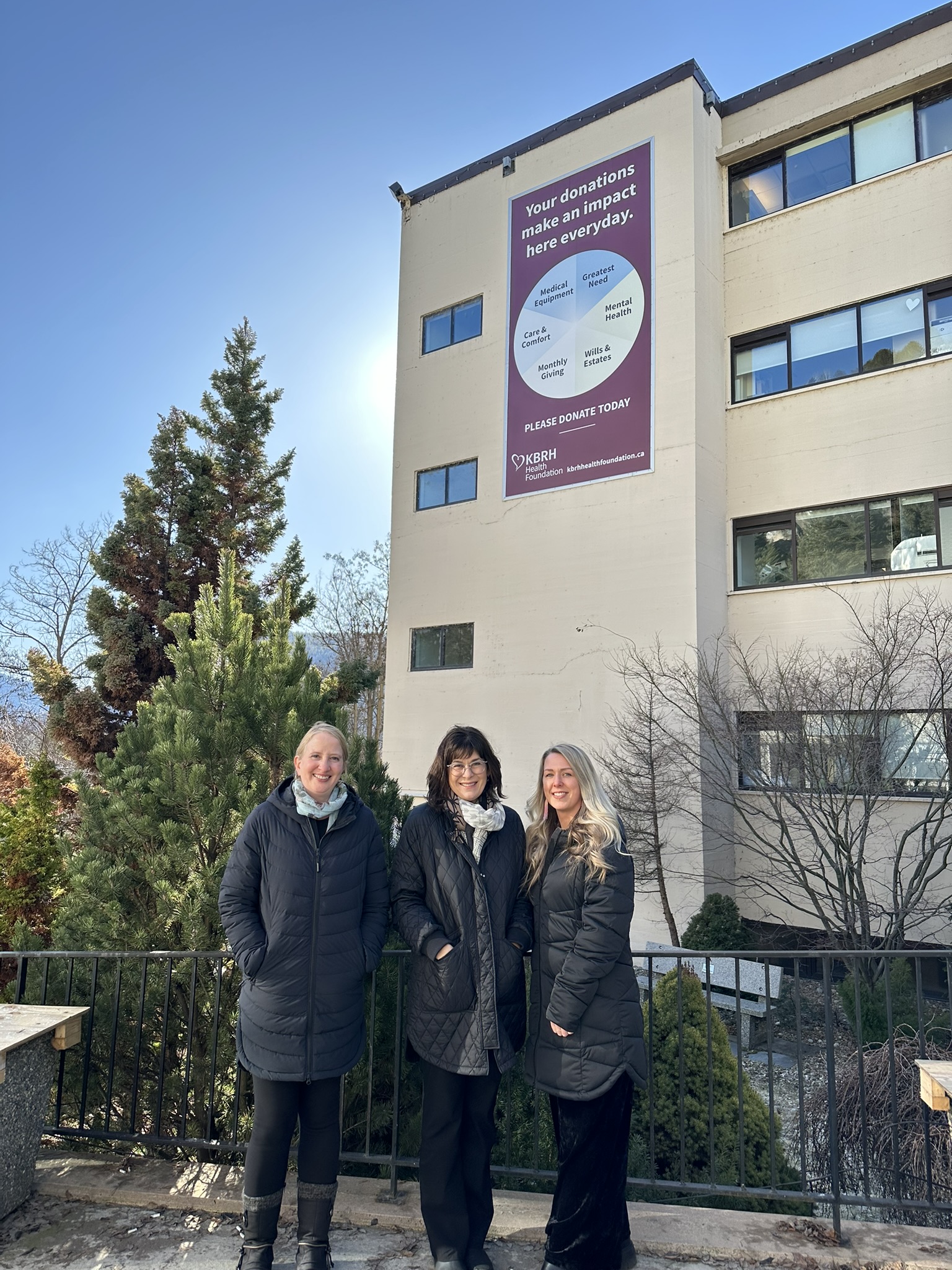 Three people pose for a picture outside with a building in the background that has a large, purple banner on it reading "Your donations make an impact here everyday."