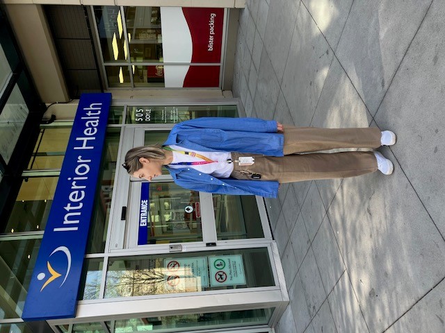 Woman with shoulder-length hair, wearing a white t-shirt, blue overshirt, and brown pants, stands in front of a building that reads "Interior Health"