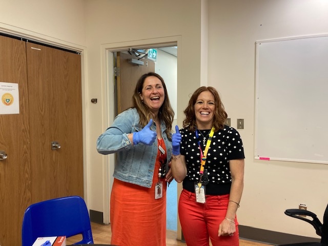 Two very enthusiastic thumbs up and big smile from one of two people standing next to each other both wearing bright colored bottoms. On the left they are wearing a blue jean jacket and the person on the right has a black polka dot shirt.