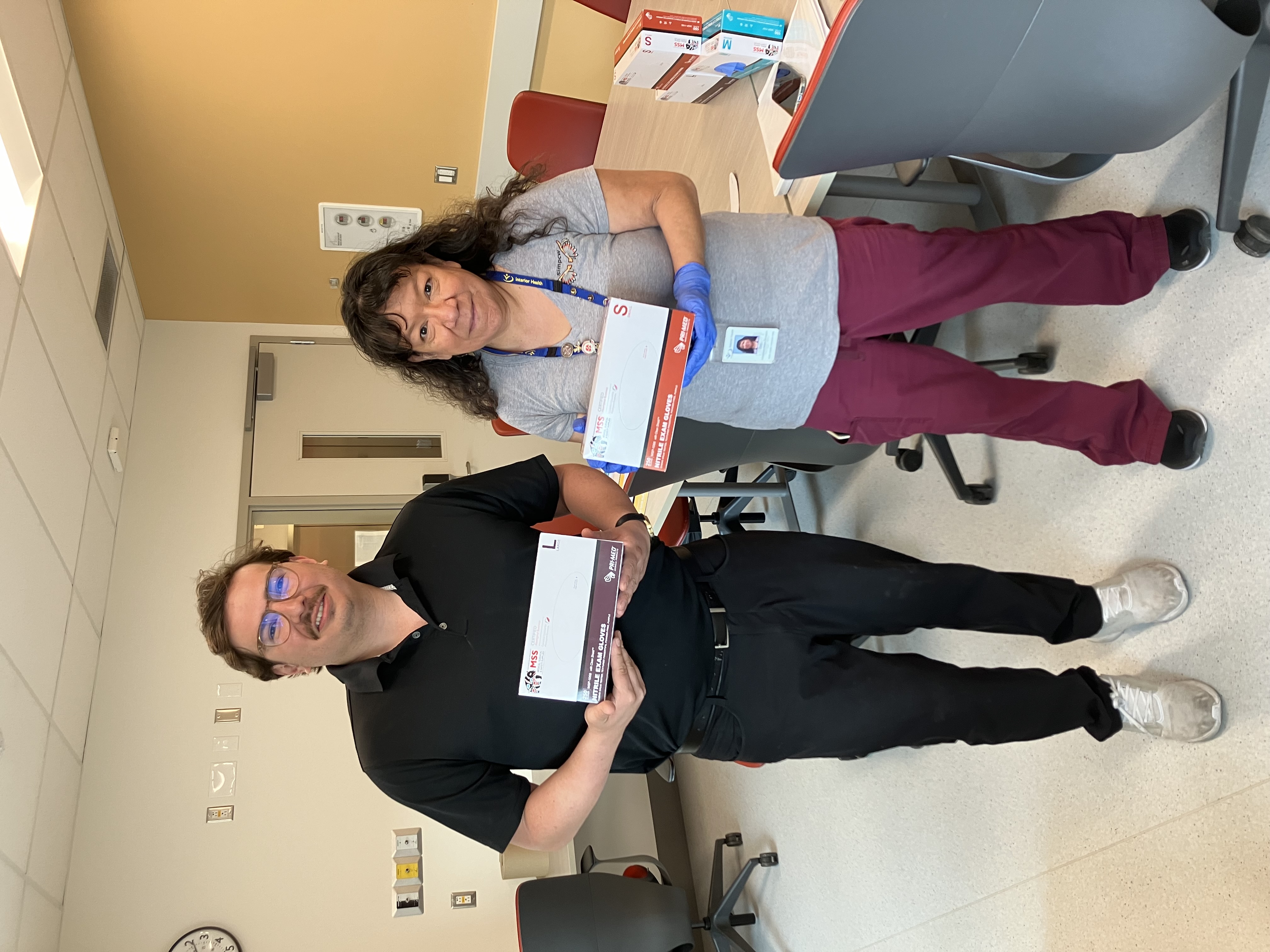 Man wearing all black stands next to a hospital staff member wearing scrubs, both are holding a box of gloves.