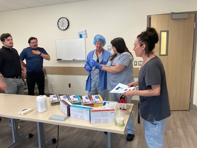 A stack of medical supplies are on a table with three hospital staff try on gloves and look at information packets while two men stand in the background.