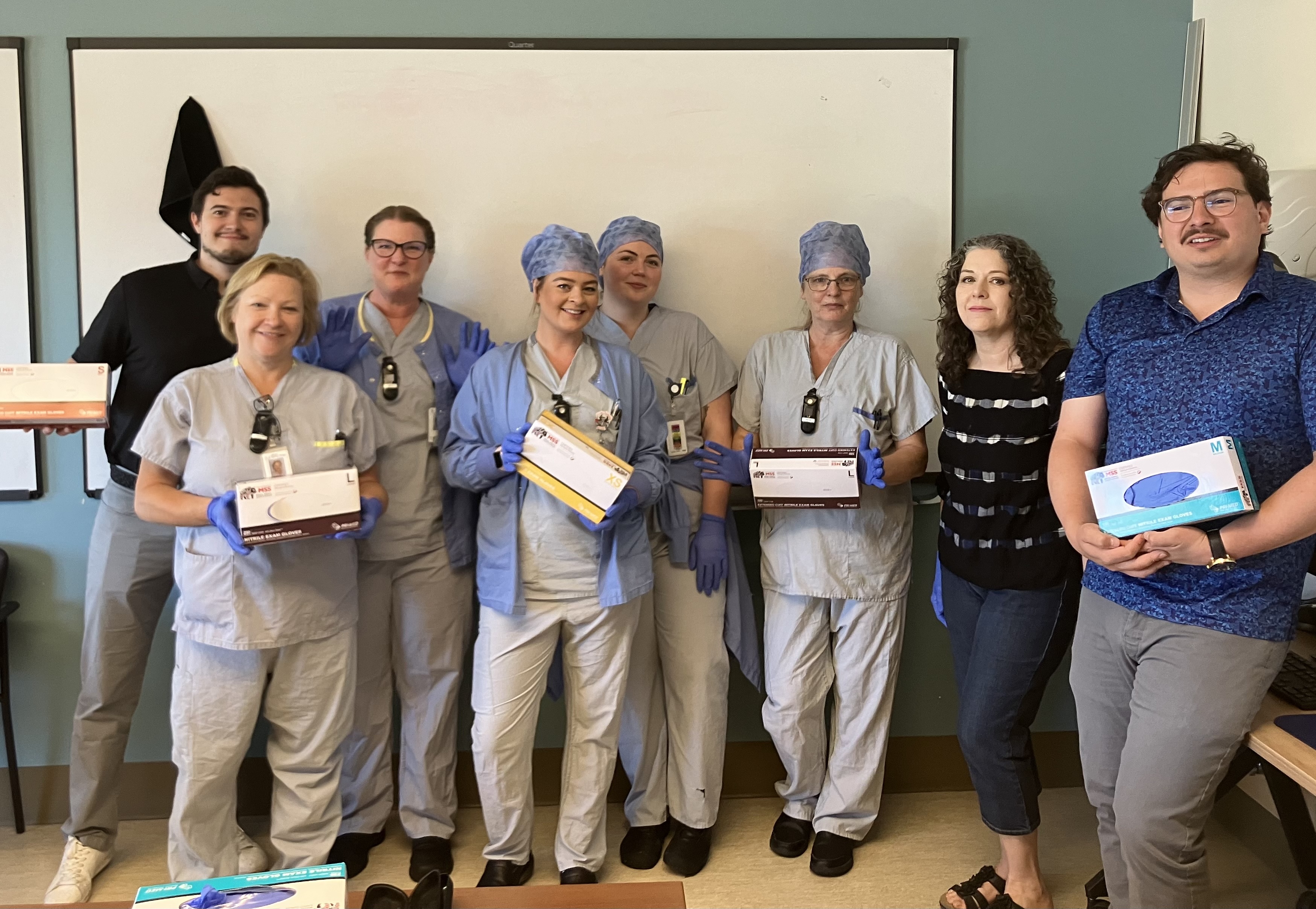 Five hospital staff wearing medical gloves holding small boxes standing between three plain clothes people also holding boxes of gloves are smiling, standing against a white board.