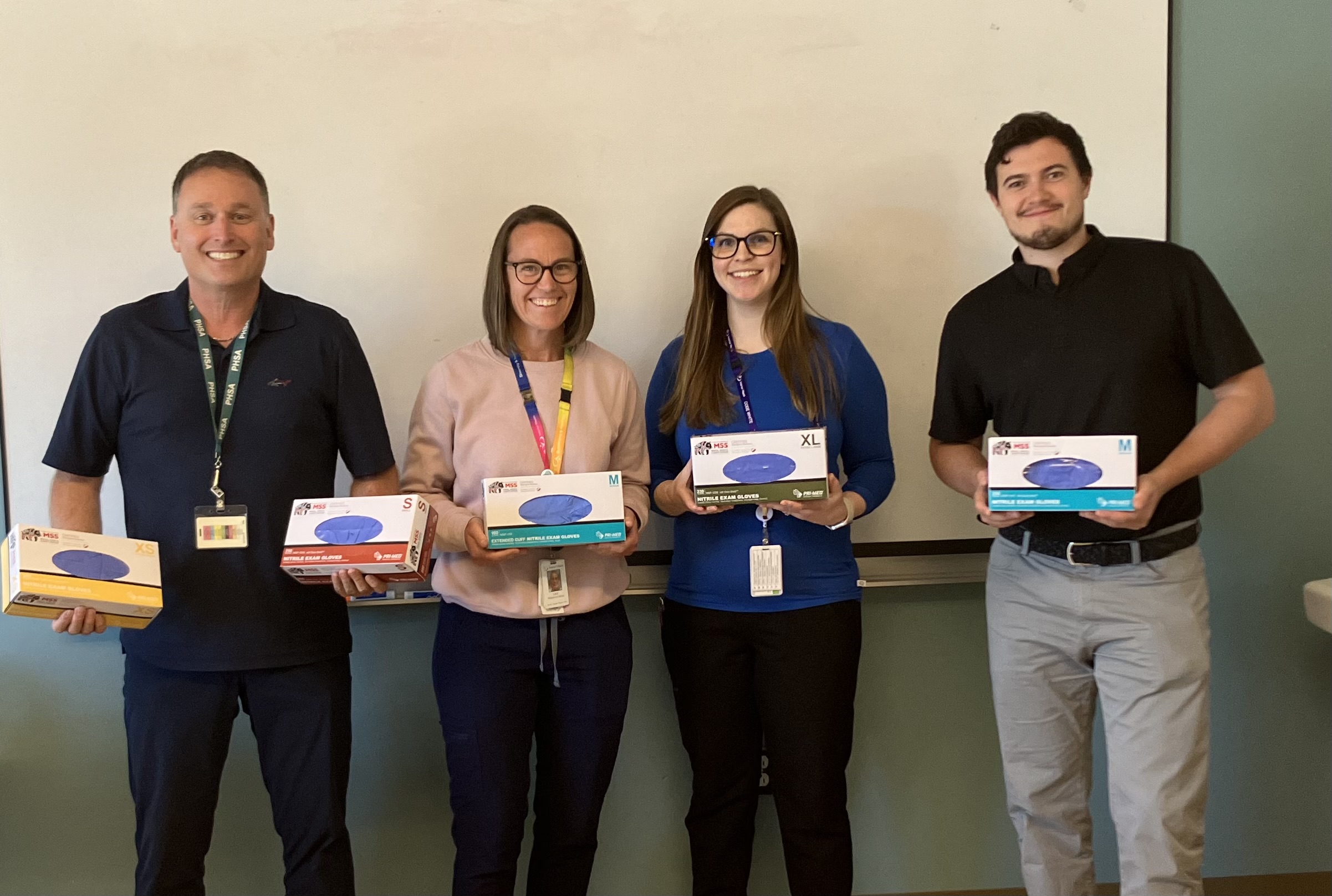 Four people standing in front of white board, all smiling, man on left is holding two boxes of gloves, four on the right are each holding one box. 