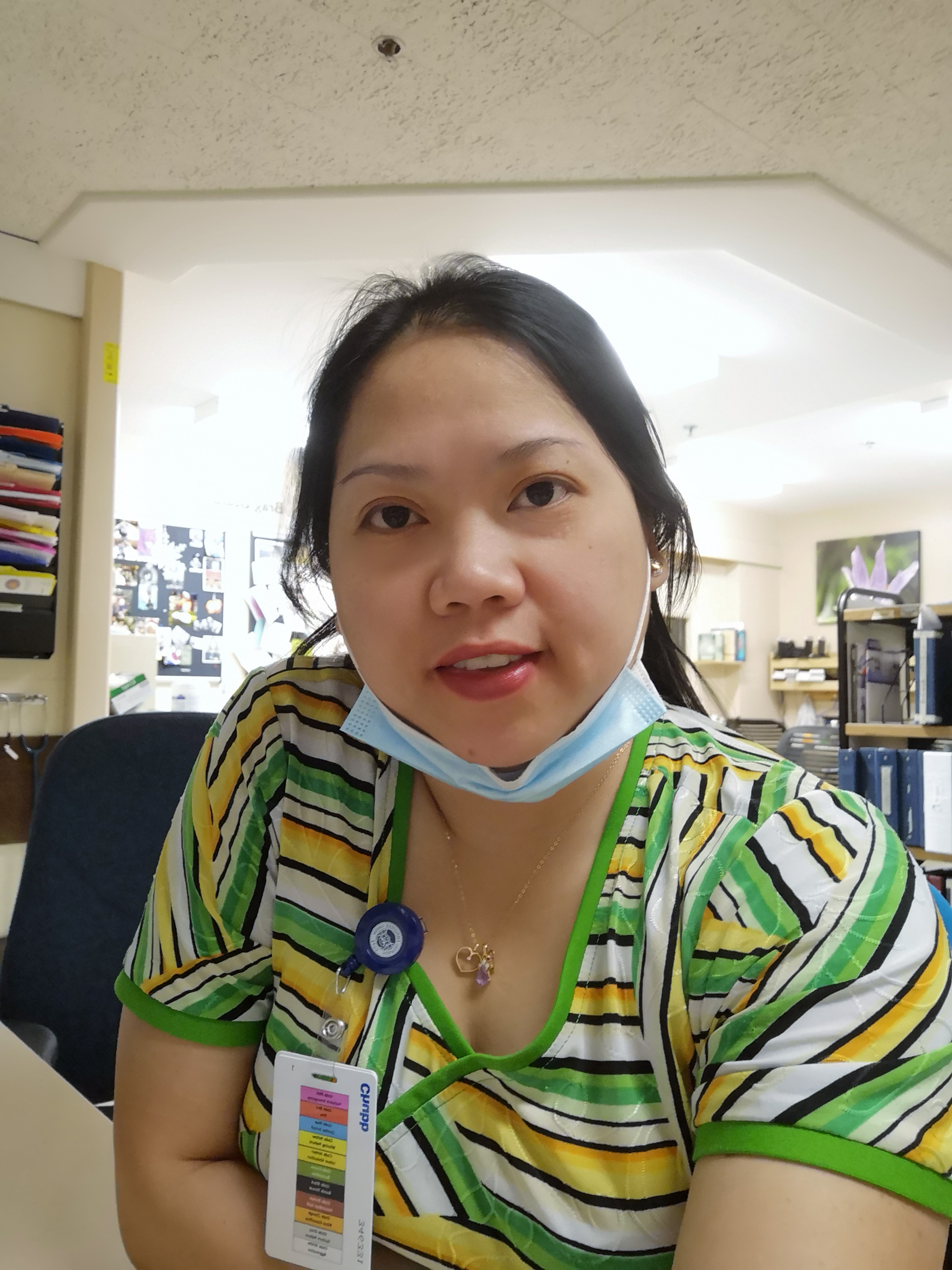 Woman facing the camera and wearing a facemask, with a green shirt while working as a care aide. 