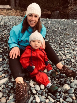 A woman with long dark hair and a cream coloured toque and a teal rain jacket sits on a rocky beach with a young boy wearing a red jumpsuit and cream coloured toque