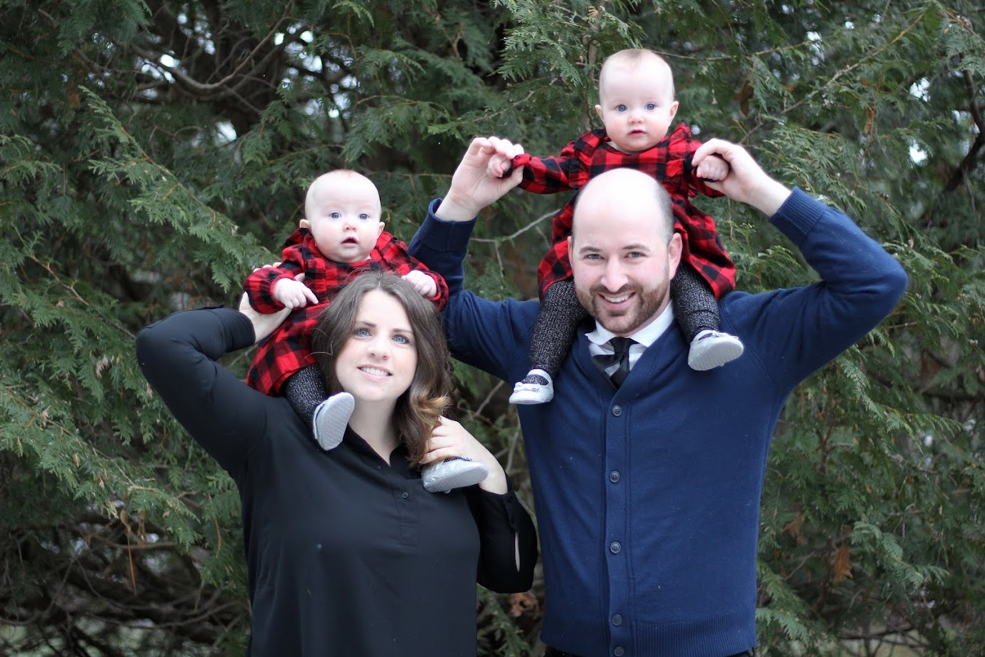 Two adults and two babies in matching plaid outfits pose for a photo outdoors, with the adults holding the babies above their shoulders.