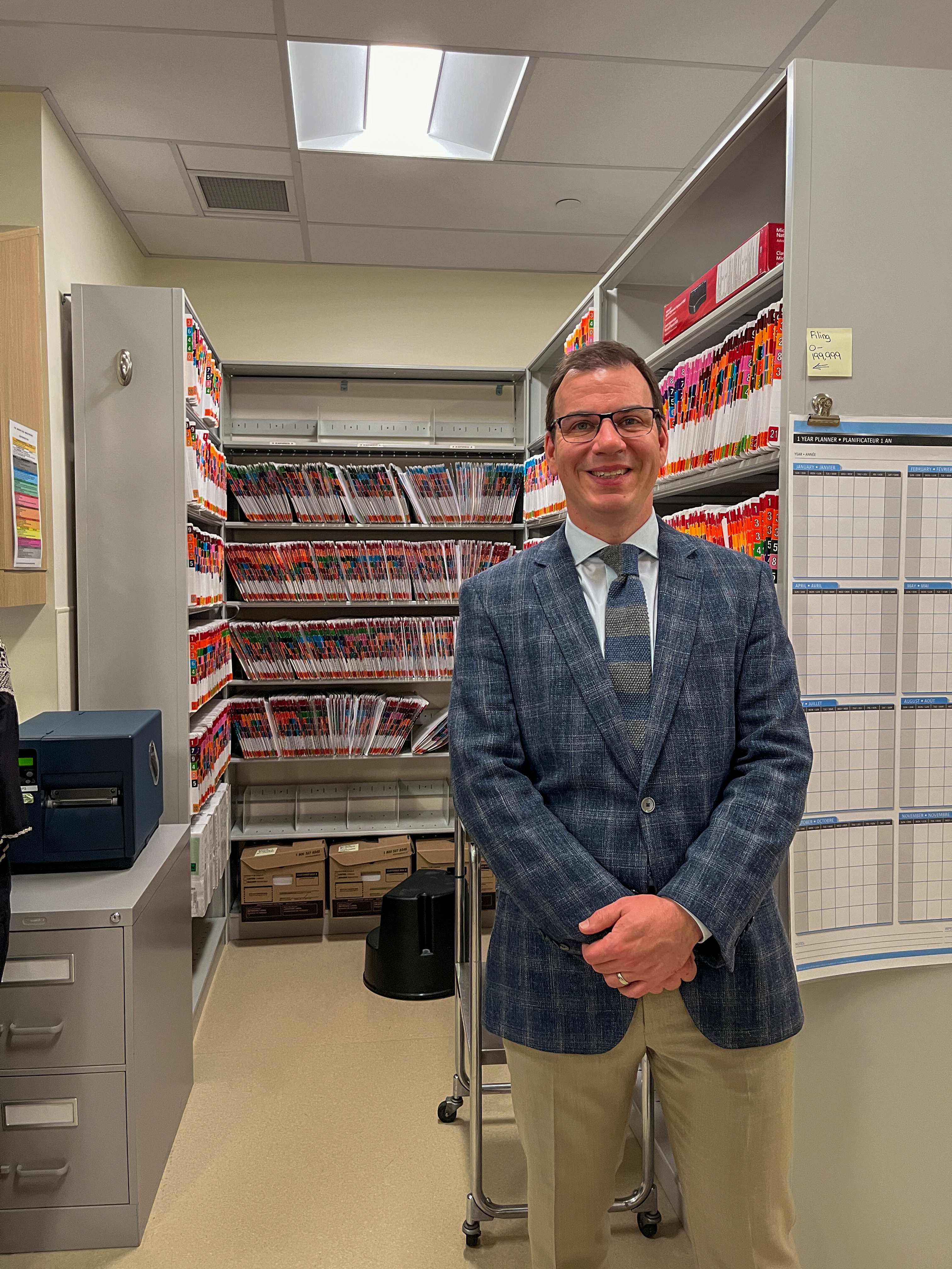 Individual in glasses and a blue checked jacket smiling in a hospital, standing in front of colourful files on shelves. The setting includes a refrigerator and a whiteboard with a grid, hinting at an organized workplace environment.