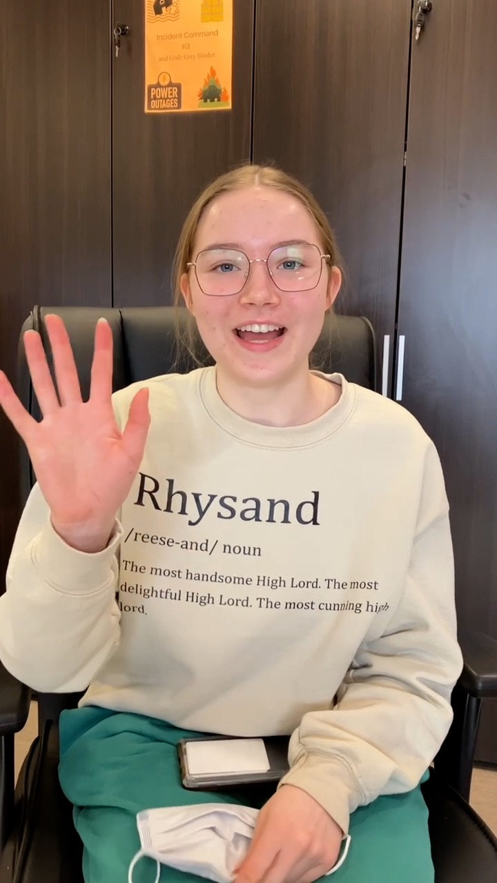 A young smiling girl with round glasses and reddish hair wearing a beige sweatshirt and green pants waves at the camera.