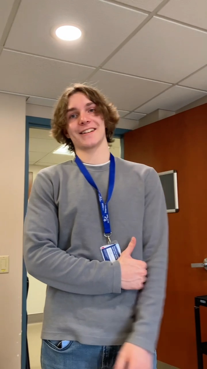 A young man with brown medium-length curly hair wearing a grey long-sleeved shirt and blue lanyard stands in front of a wood door in a classroom. 