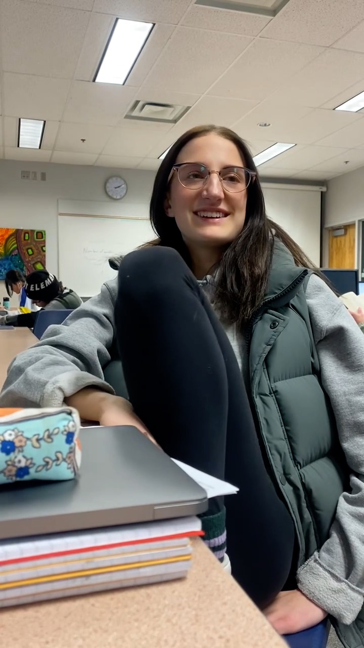 A smiling girl with long dark hair and glasses wearing a green puffy vest and grey sweatshirt sits at a desk in a school classroom.