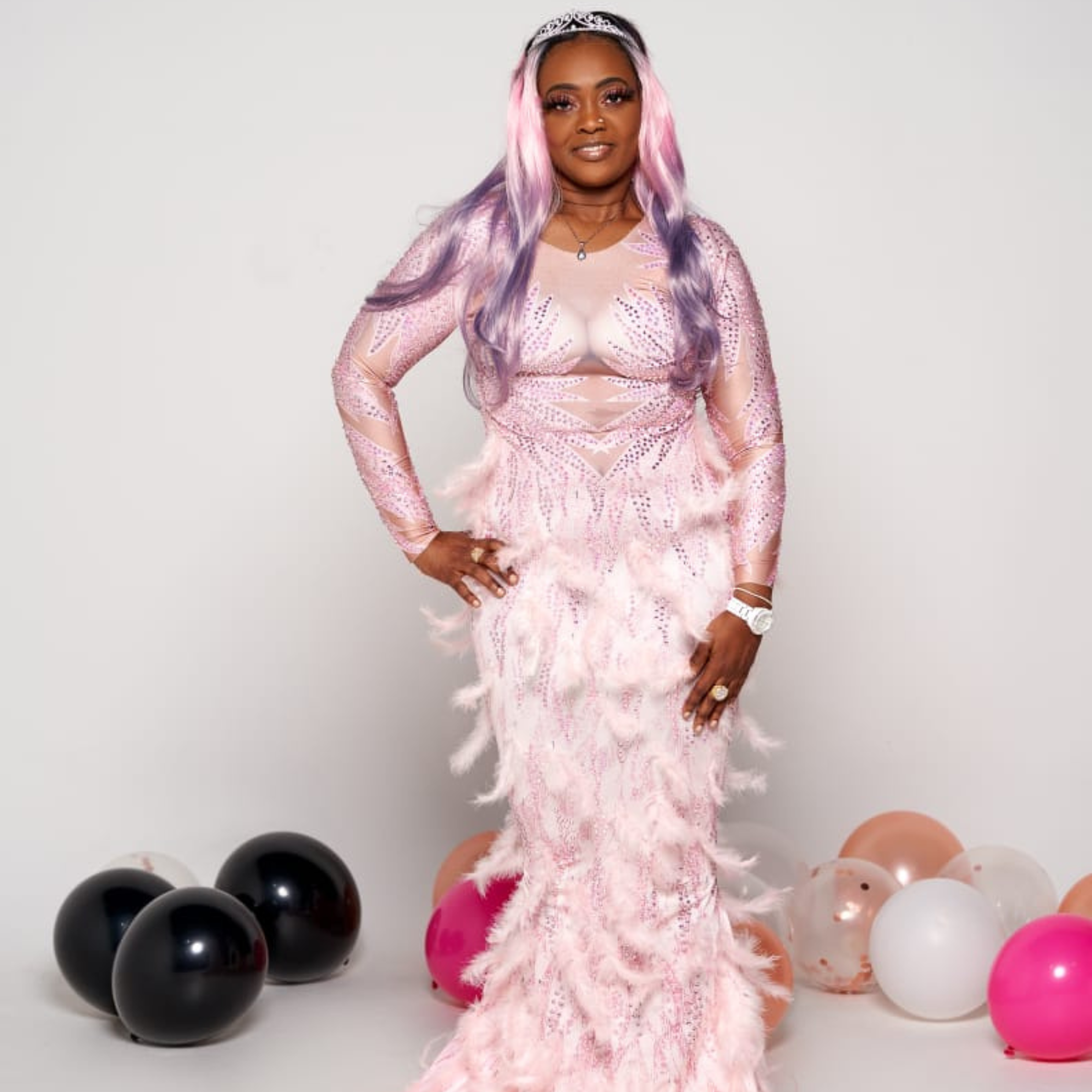 A smiling woman with blonde, pink and purple hair wearing a pink feathered floor length gown standing in front of balloons that are laying on the floor. 