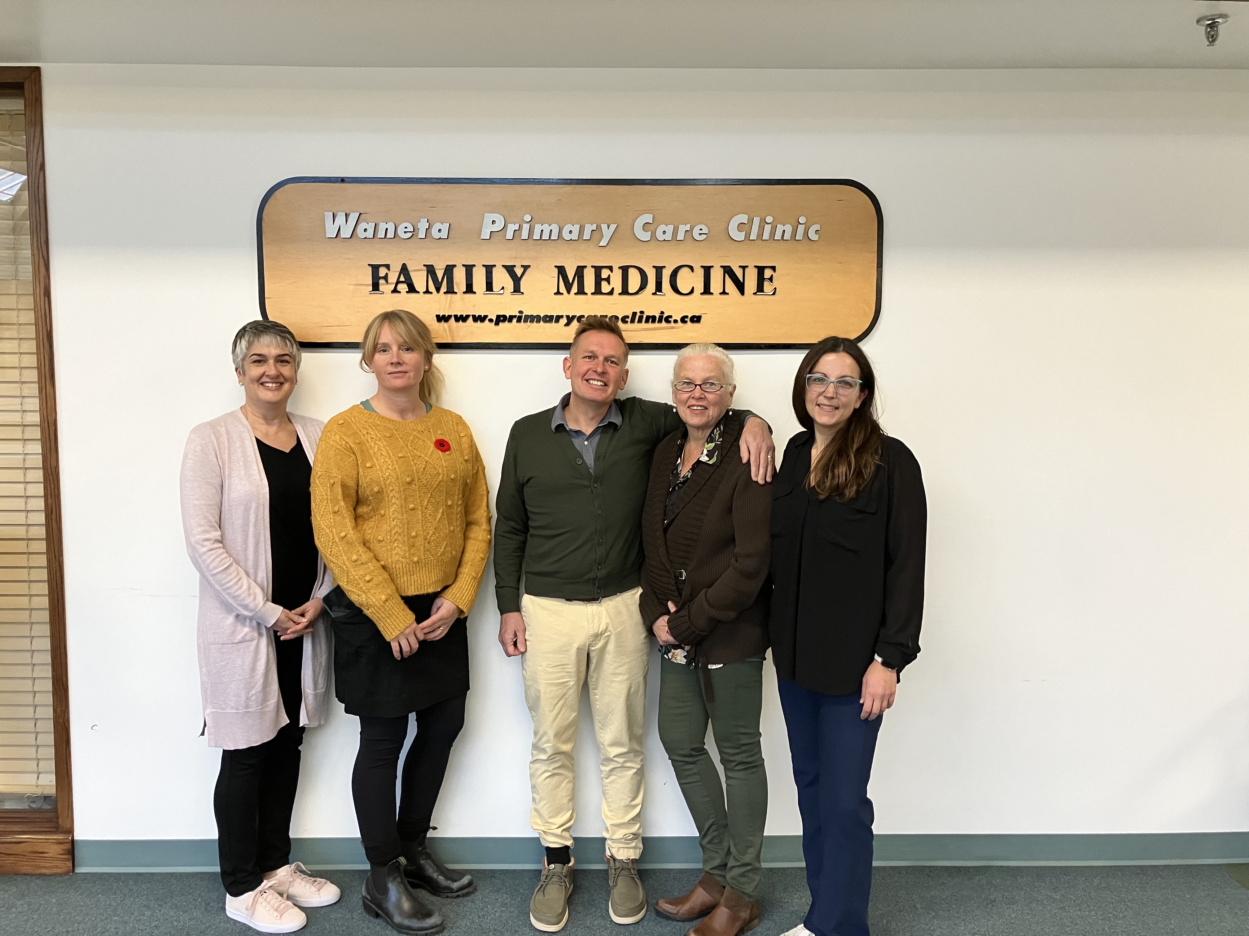 A man standing in the middle of four women, two on each side, all smiling, posing for a photo in front of a sign that reads, Waneta Primary Care Clinic.