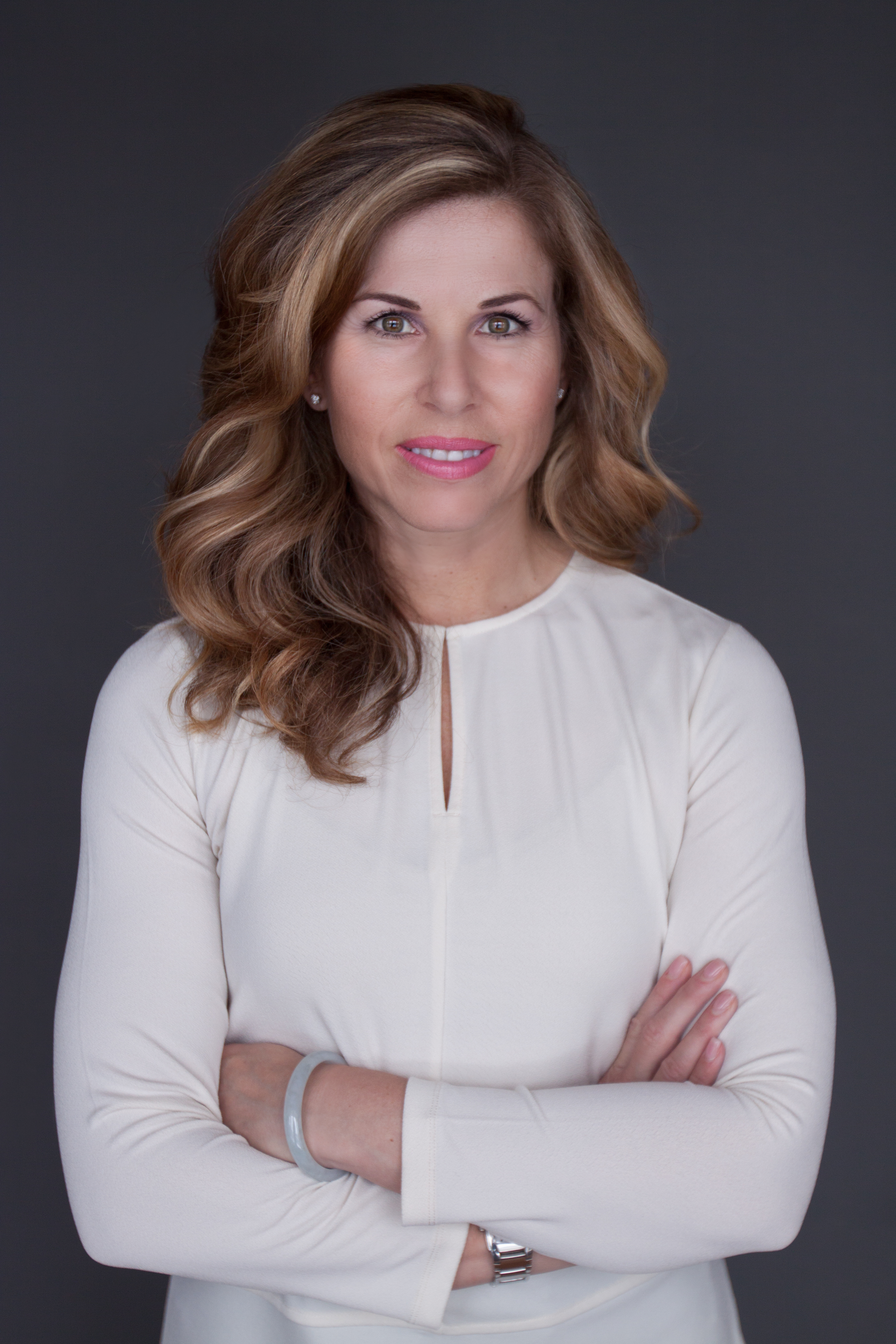 A woman with dirty blonde, shoulder length, curled hair wearing a white blouse and pink lipstick, standing with her arms folded in front of her, smiling and posing for the camera.