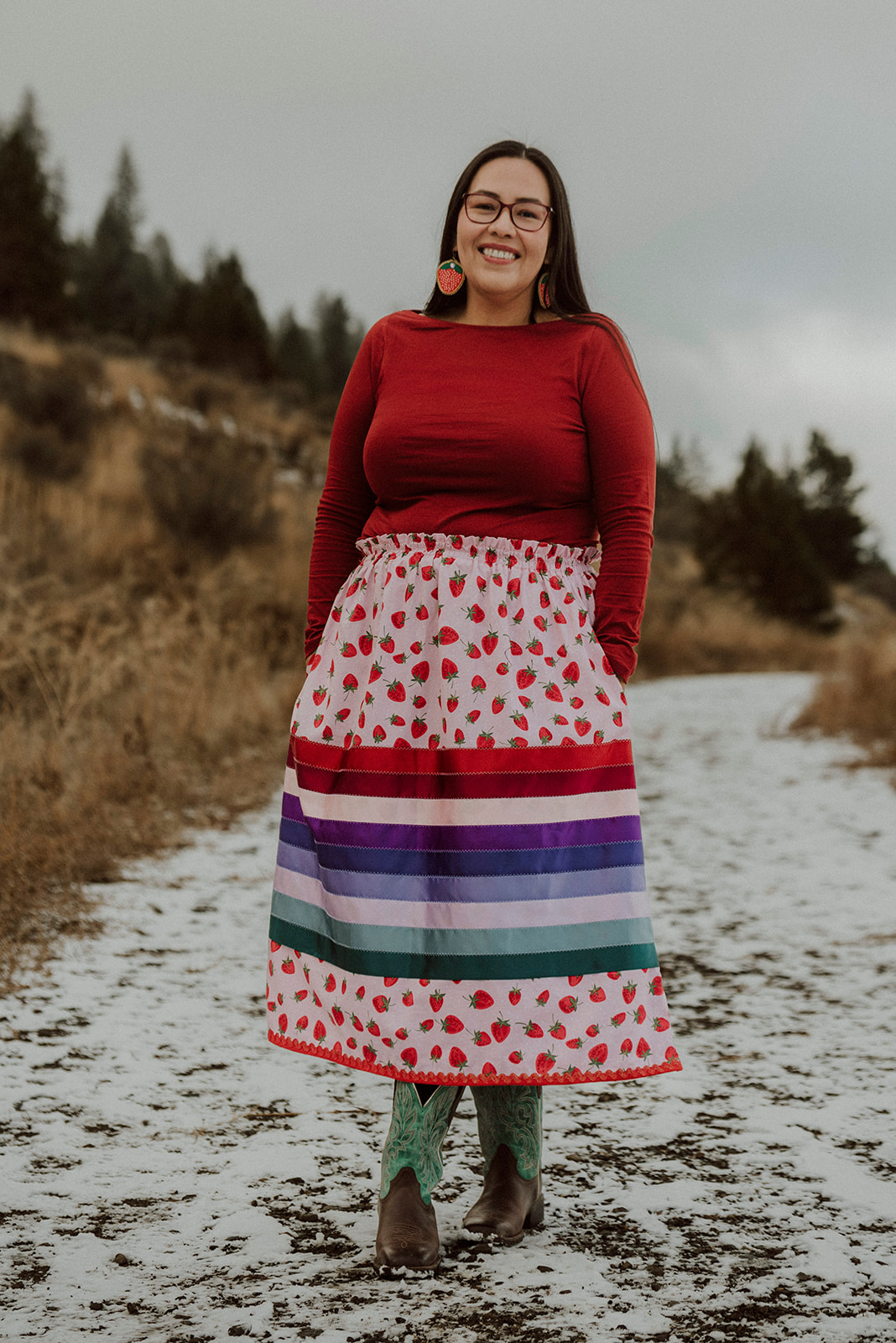 A woman with long dark hair, wearing strawberry earrings, dark rimmed glasses, a long-sleeved red shirt and a strawberry ribbon skirt is standing outdoors on a snowy path amongst a wooded area, smiling for the camera.