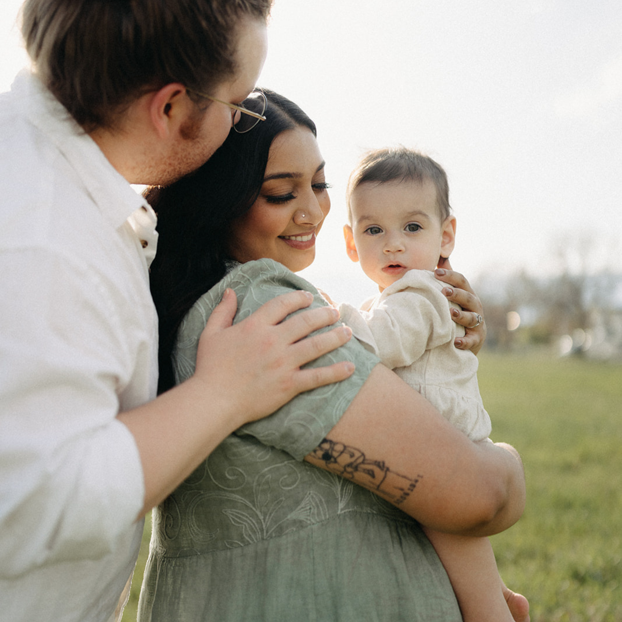 A family, with a dad kissing a pregnant mom’s head while holding their baby, poses outside in the summer. 