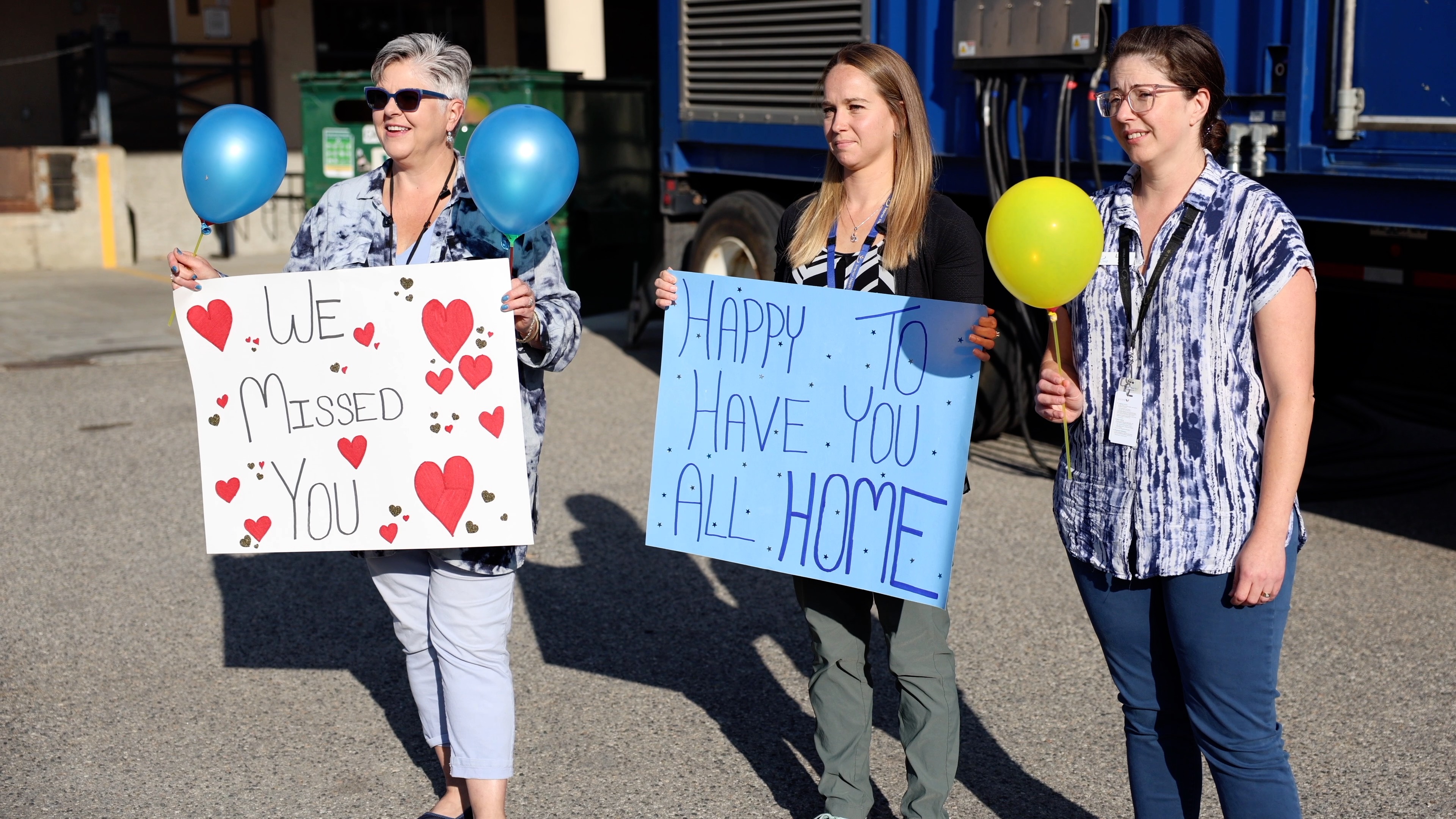 Three people stand outside holding balloons and two signs, one with hearts that says we missed you, and another with stars that says happy to have you all home.