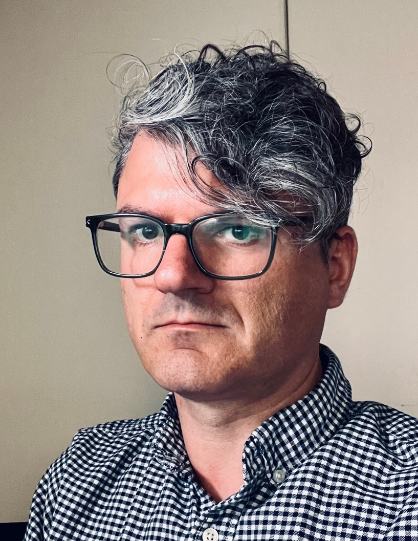 A person with curly salt and pepper coloured hair and black square glasses wearing a black and white checkered shirt looks unsmiling at the camera.