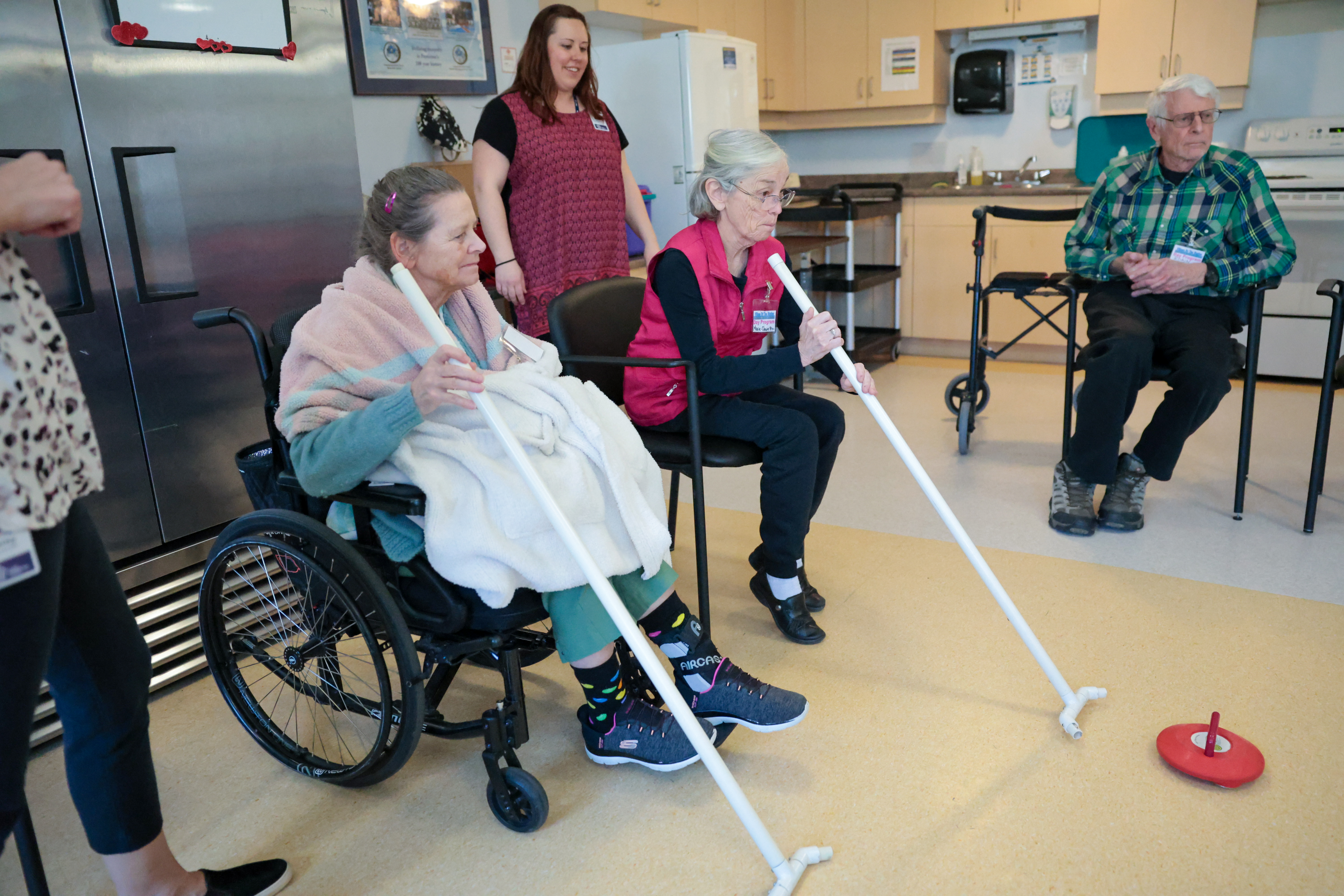 Two women, one with grey hair, a pink shawl and teal pants and one with grey hair, glasses and a pink vest and black pants hold curling sticks pointed at a curling rock as a few people look on