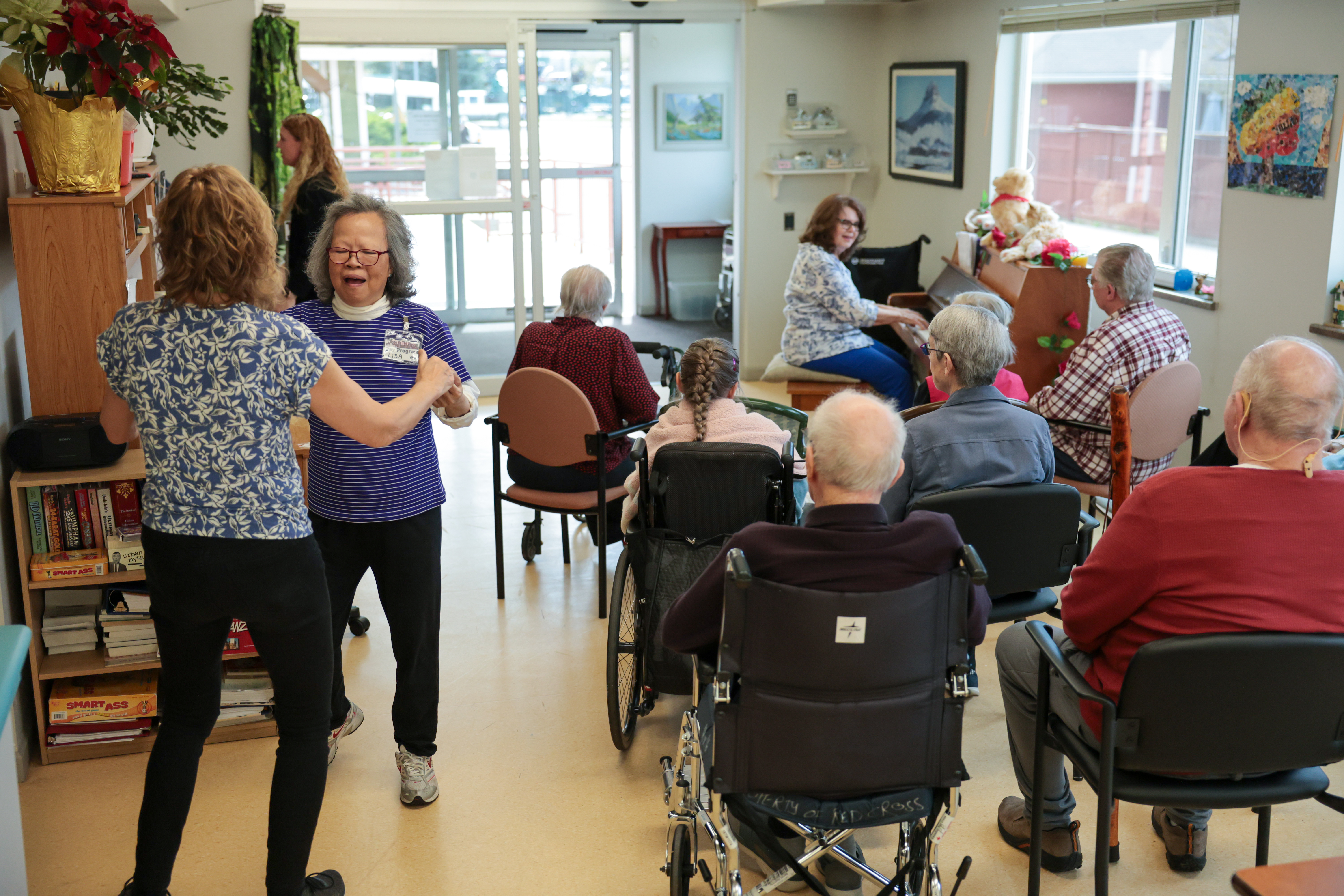 A large group of older people sitting in chairs watches a woman in a white and blue shirt and blue pants play the piano and sing